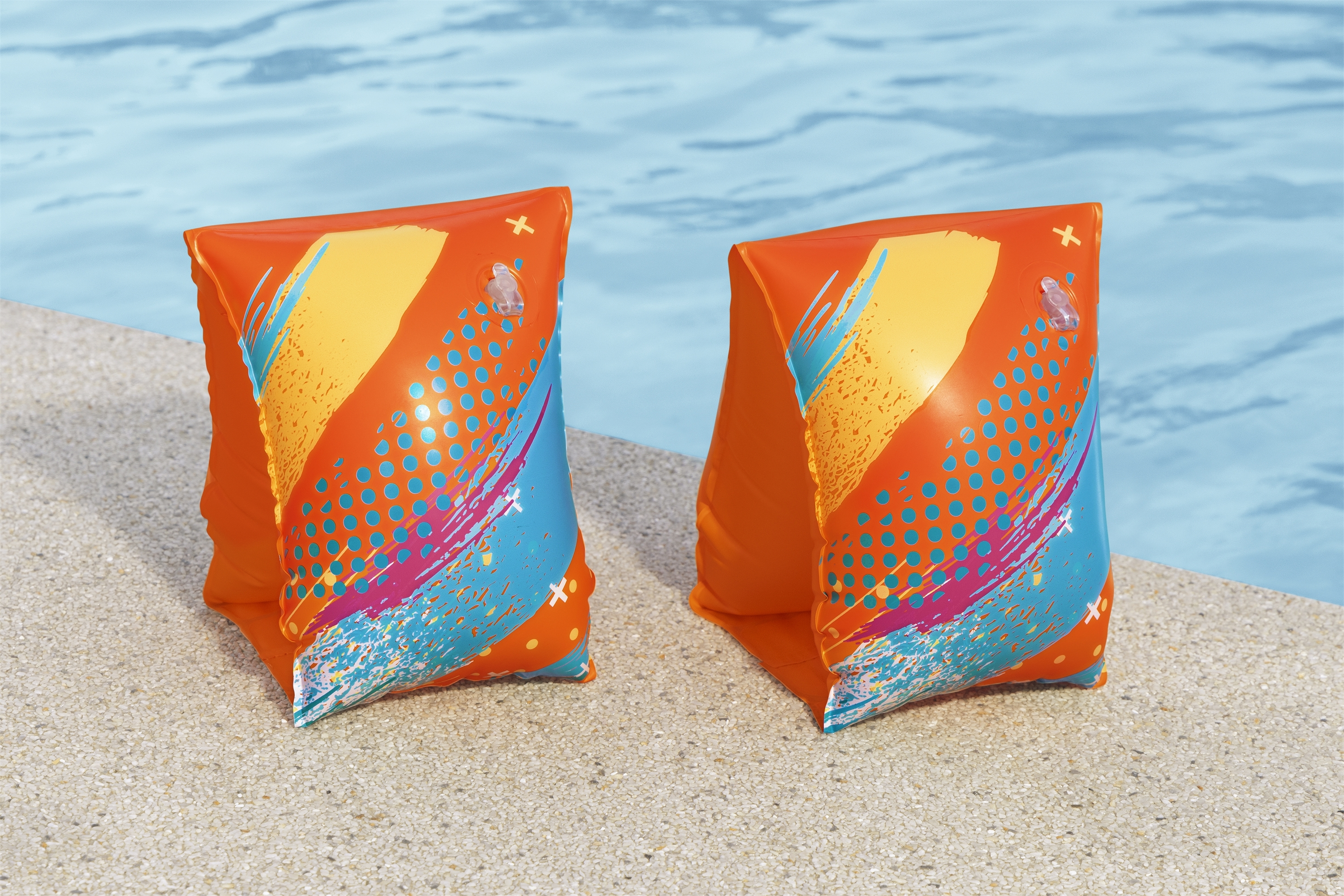 Zwei orangefarbene Armbänder mit bunten Designs liegen auf einem Pooldeck neben einem Pool mit blauem Wasser.