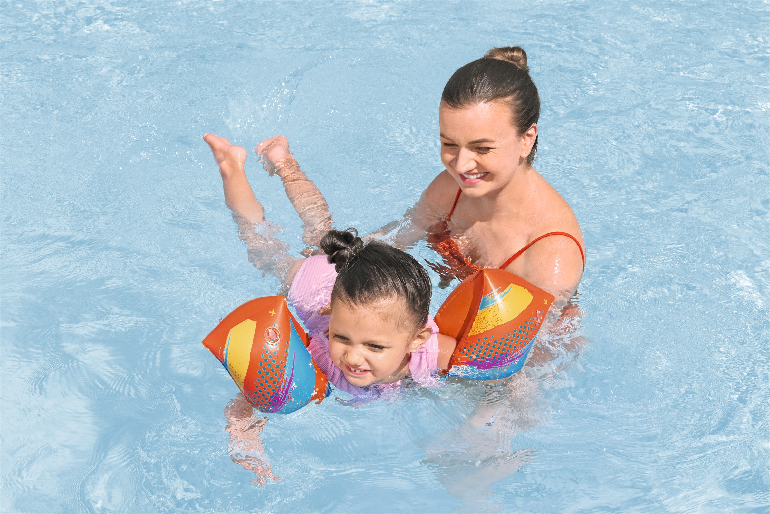 Eine Frau und ein Kind sind in einem Pool. Das Kind trägt Schwimmflügel. Beide lächeln.