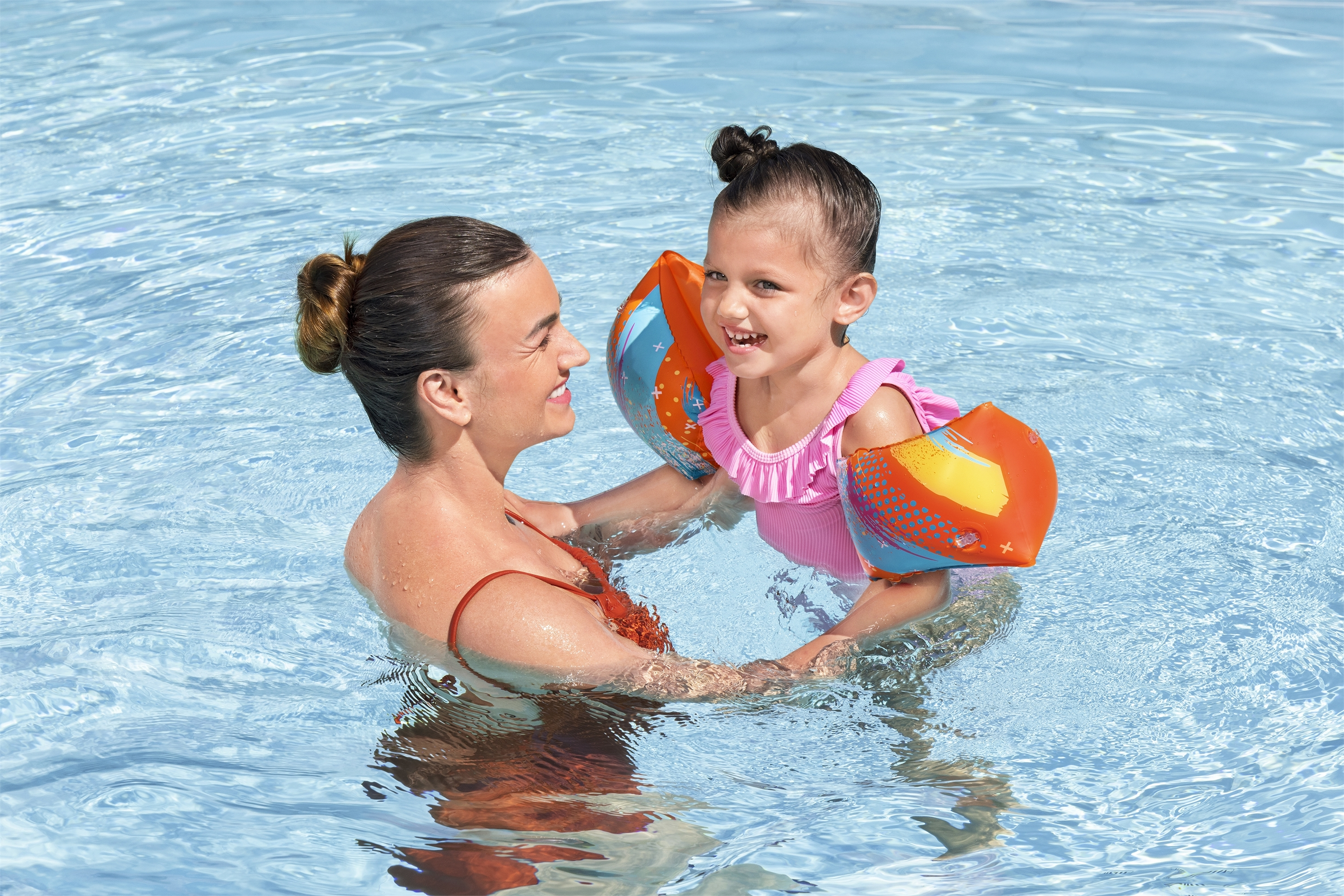 Eine Frau und ein Kind sind in einem Pool. Das Kind trägt Schwimmflügel, lächelt und wird von der Frau gehalten.