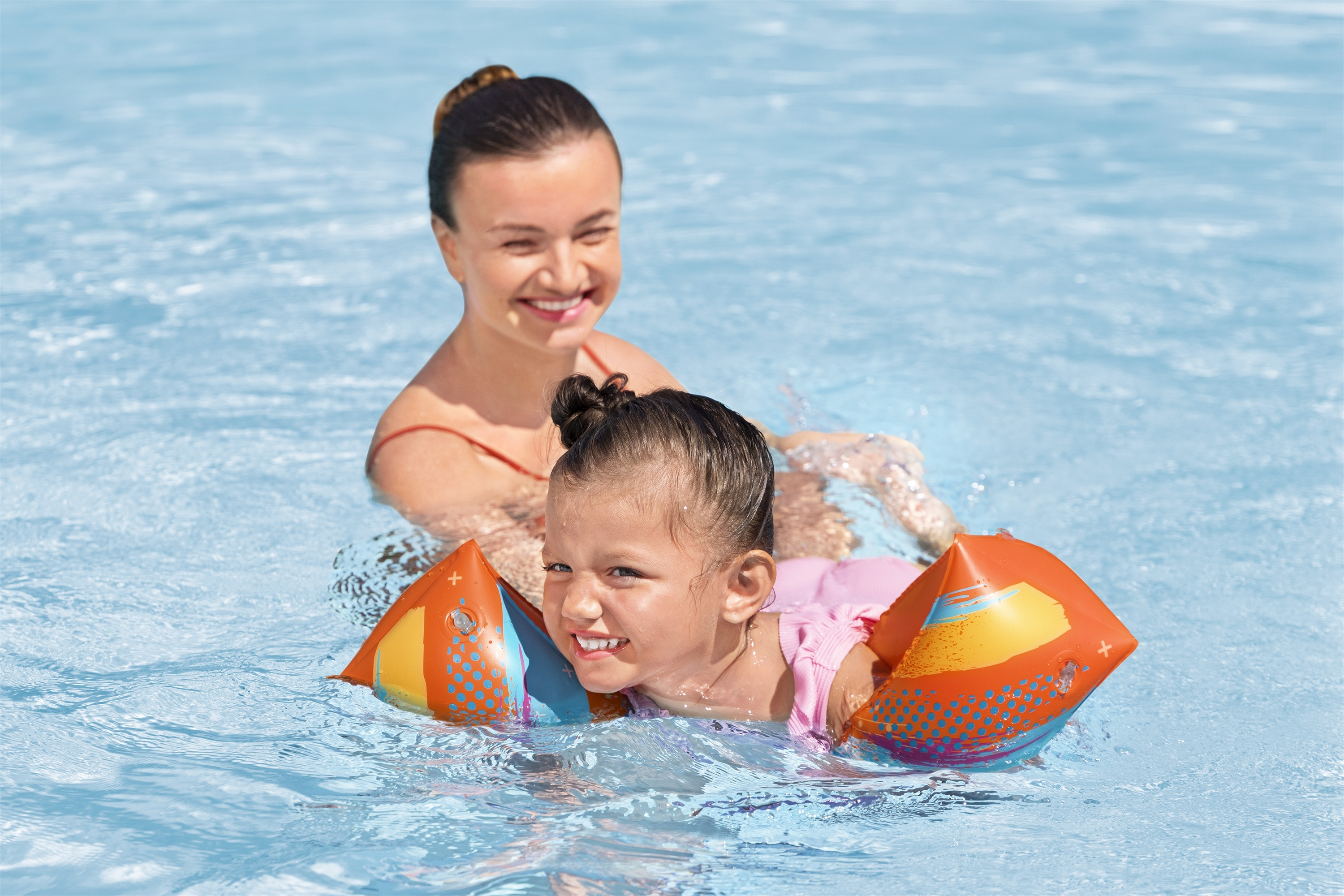 Ein Mädchen im Pool mit Schwimmflügeln lächelt. Eine erwachsene Frau lächelt hinter ihr.