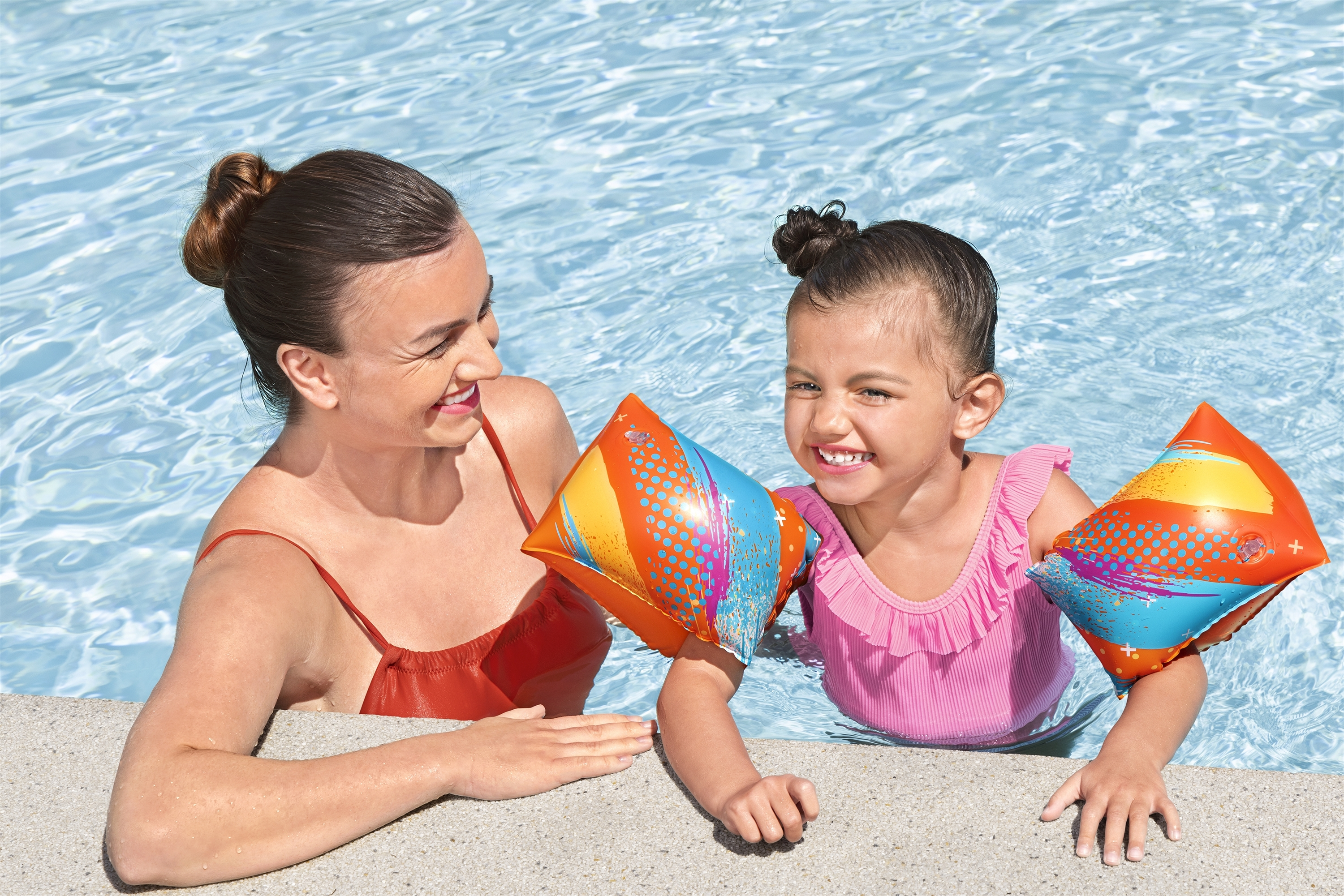 Eine Frau und ein Mädchen sind in einem Pool. Das Mädchen trägt Schwimmflügel und einen rosa Badeanzug. Beide lächeln.