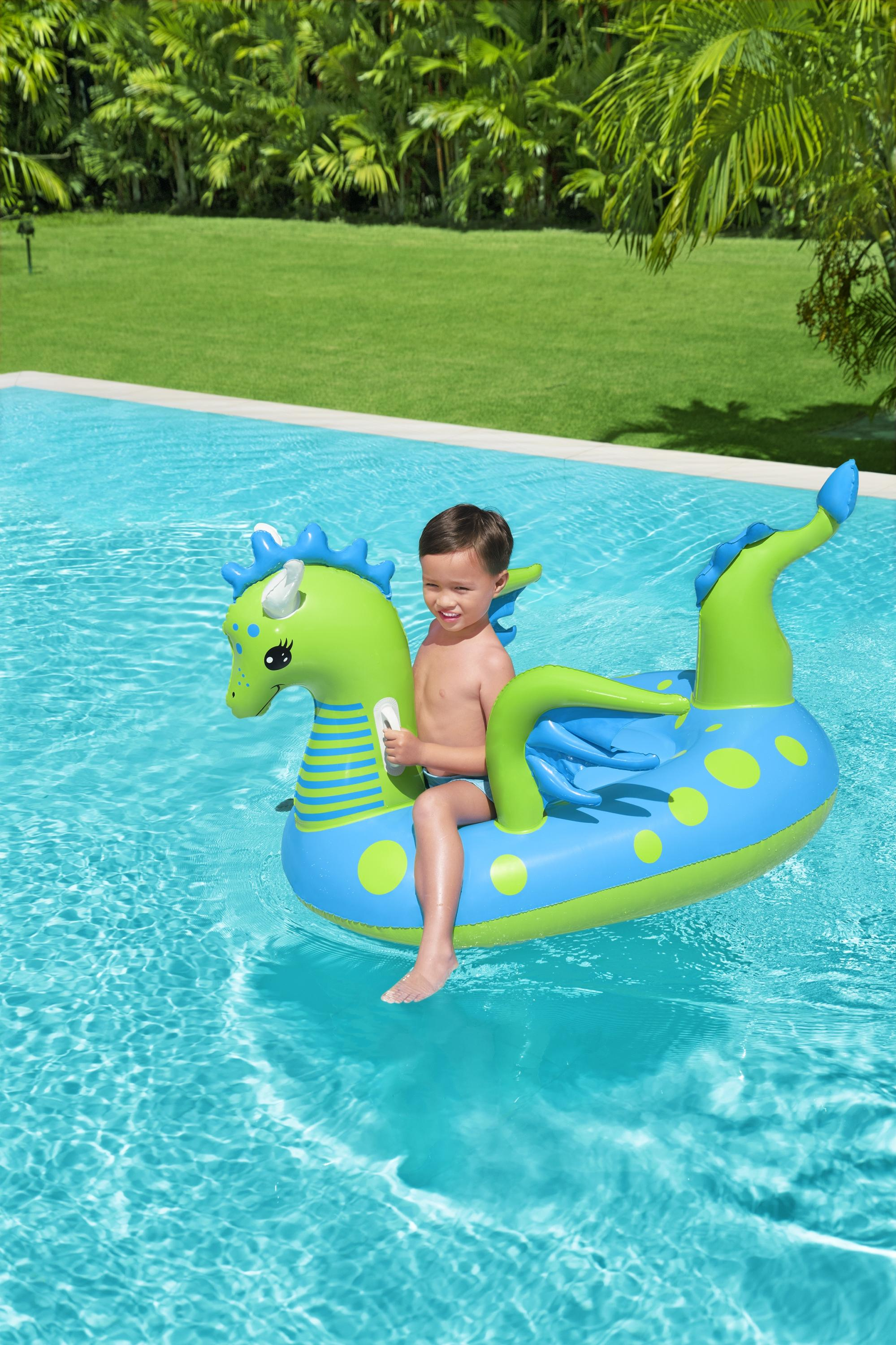 Un ragazzo cavalca un galleggiante a forma di drago in una piscina. Erba verde e alberi sullo sfondo.
