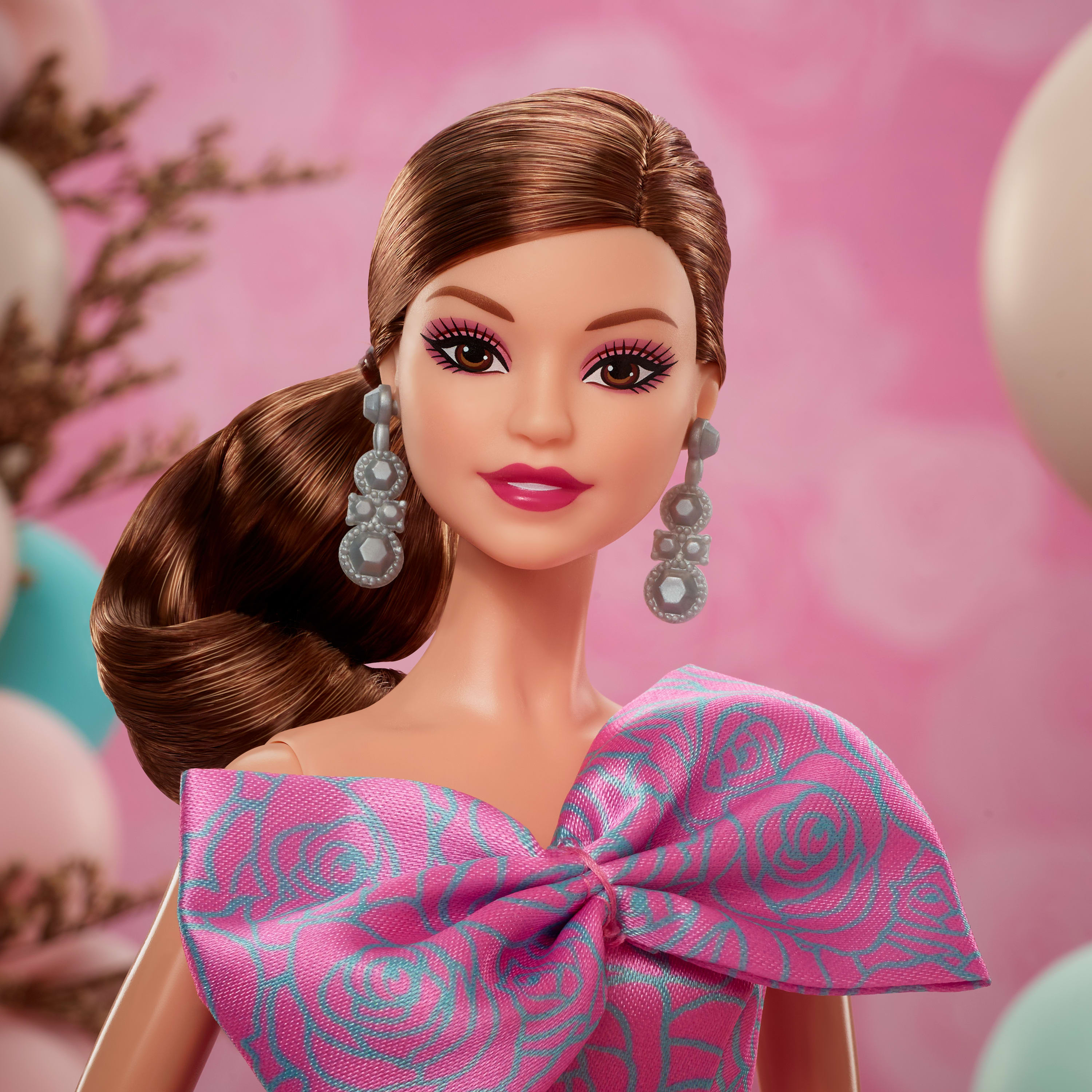 Eine Barbie-Puppe mit braunen Haaren und einem rosa Kleid. Rosa und türkisfarbener Hintergrund mit Luftballons.