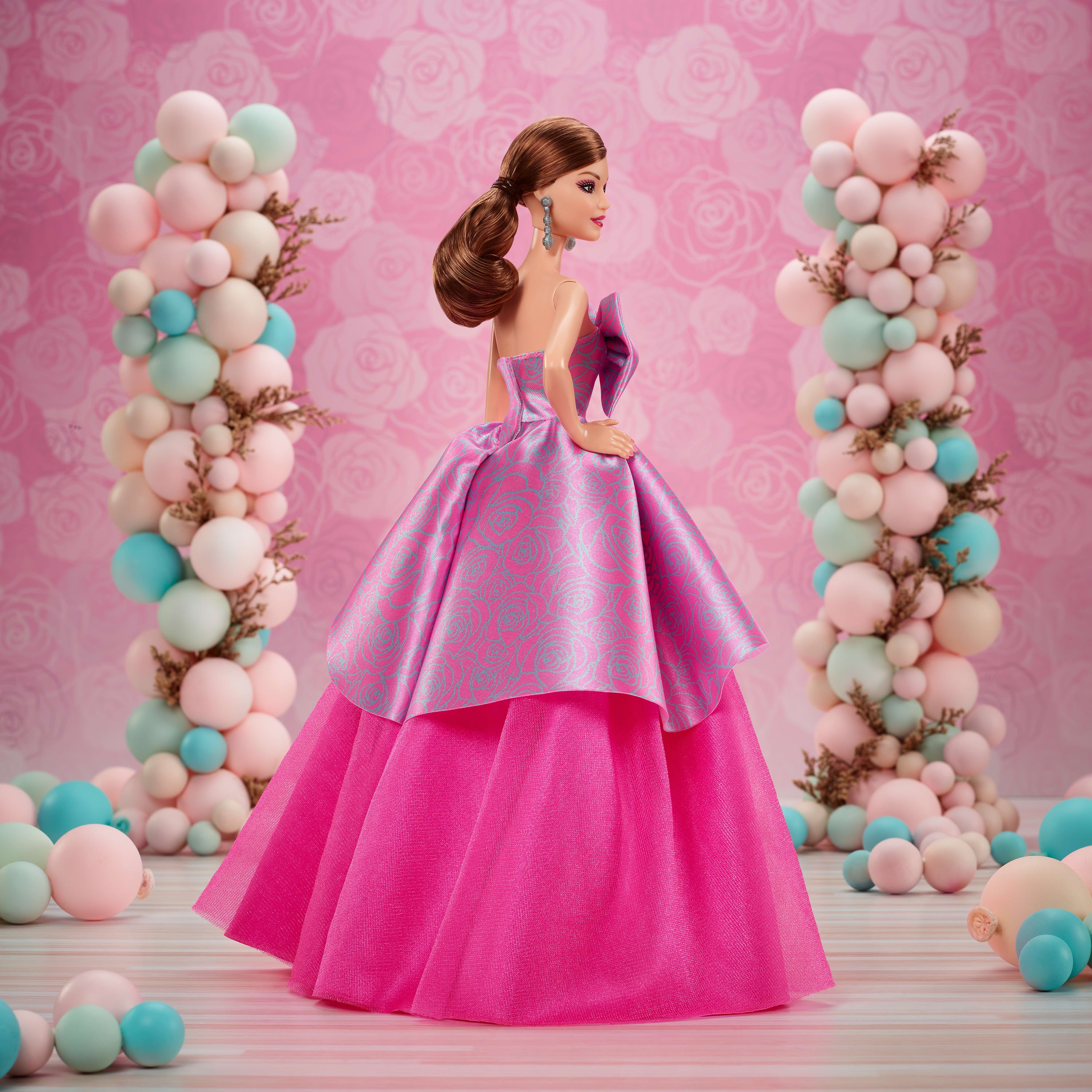 Eine Puppe in einem rosa Kleid steht neben Ballonsäulen. Der Hintergrund hat ein rosa Rosenmuster.