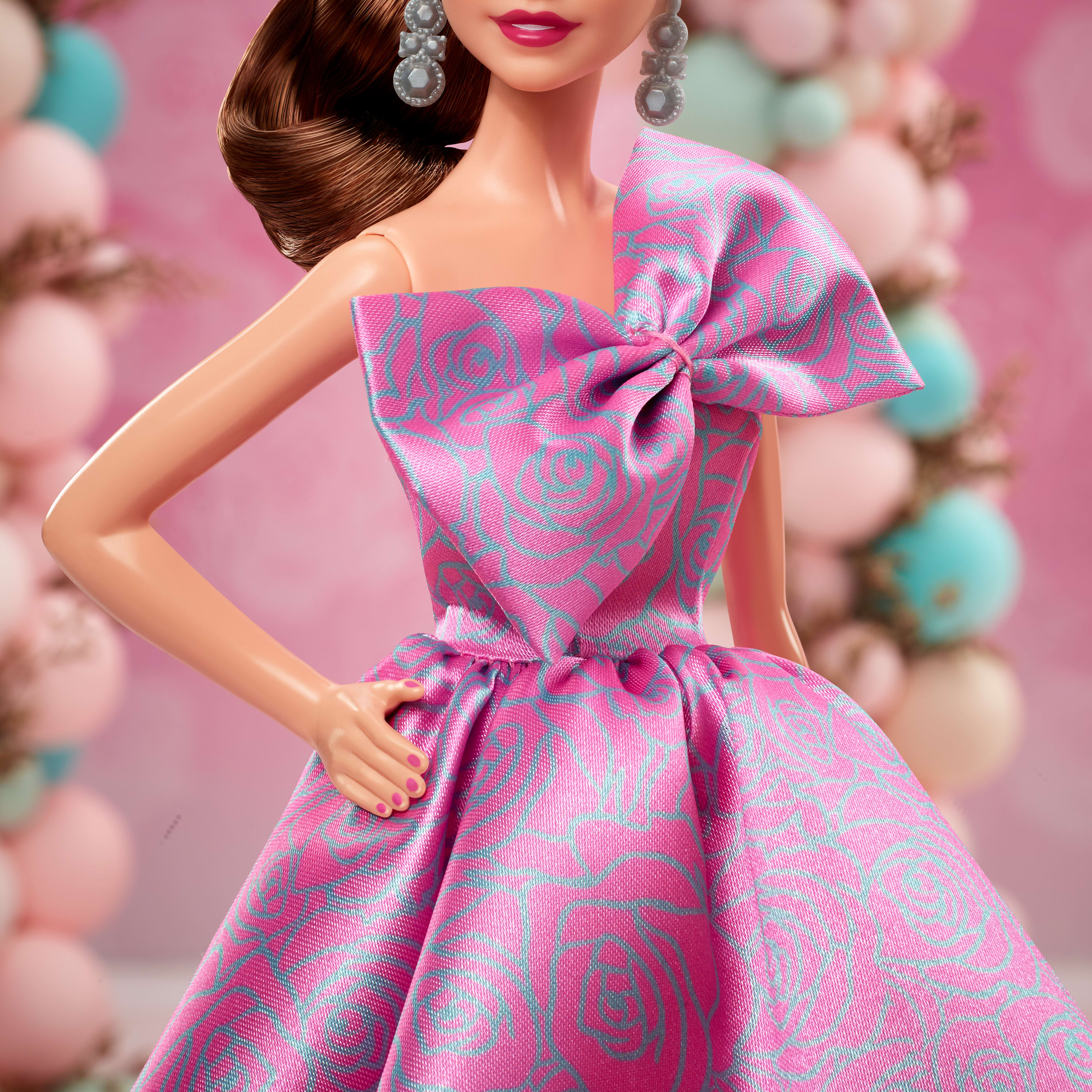 Eine Barbie-Puppe in einem pinkfarbenen Kleid mit blauem Blumenmuster. Rosa und blaue Luftballons sind im Hintergrund.