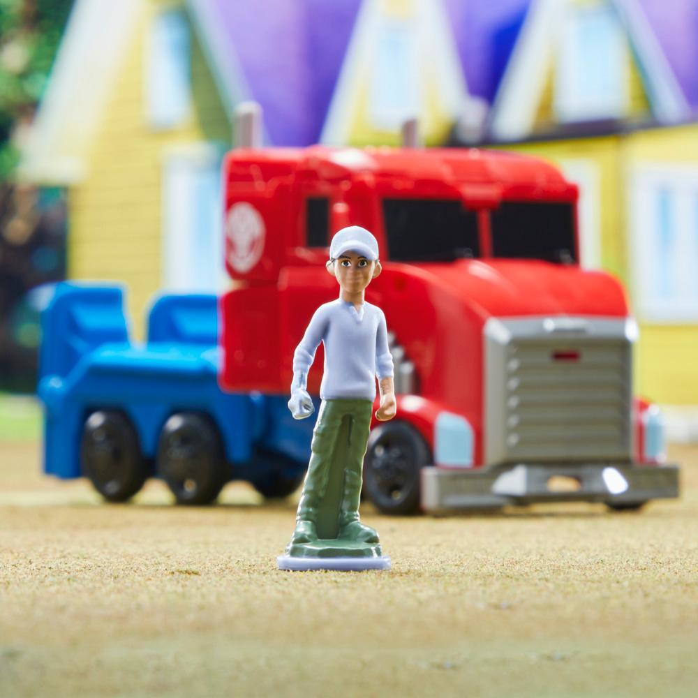 Zabawkowa figurka osoby stoi przed czerwoną ciężarówką i niebieską przyczepą. W tle żółty dom i fioletowe niebo.