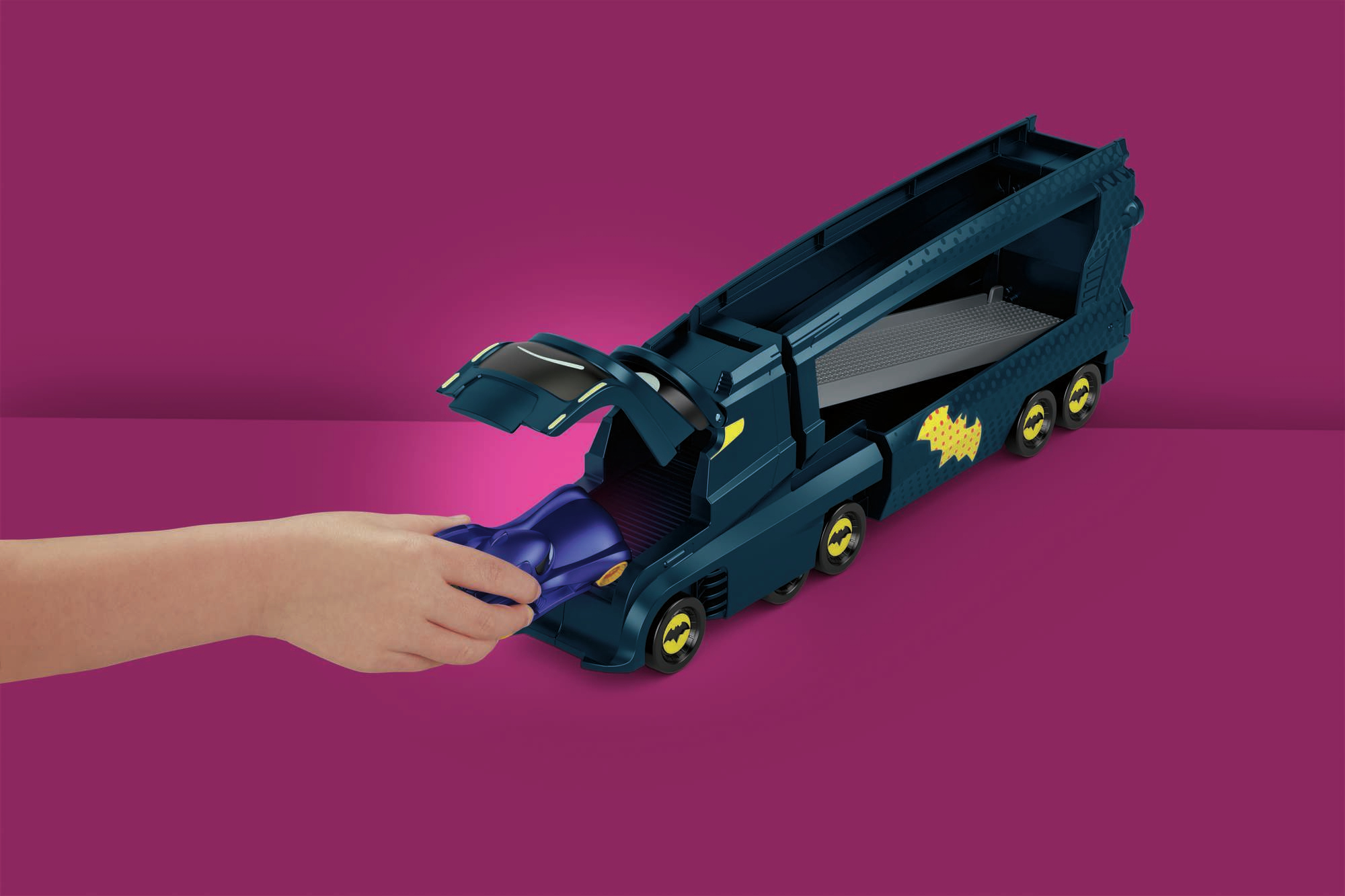 Ręka umieszcza niebieski samochodzik w ciemnoniebieskiej ciężarówce Batmana na fioletowym tle.