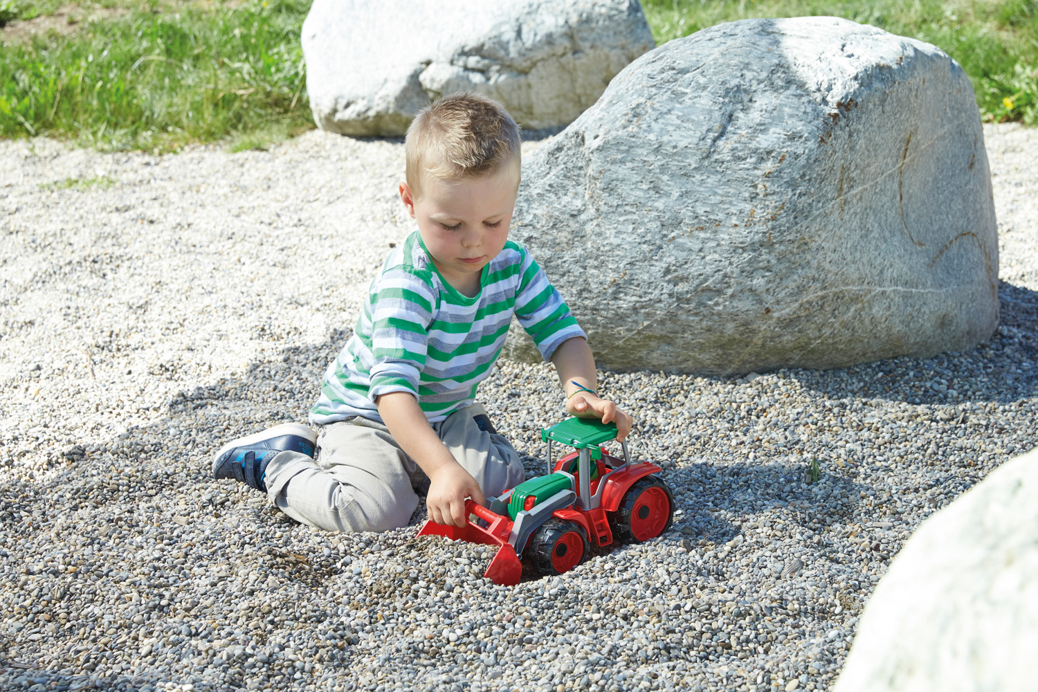 Un niño juega con un tractor de juguete rojo y verde sobre grava. Grandes rocas y hierba en el fondo. El niño lleva rayas verdes.