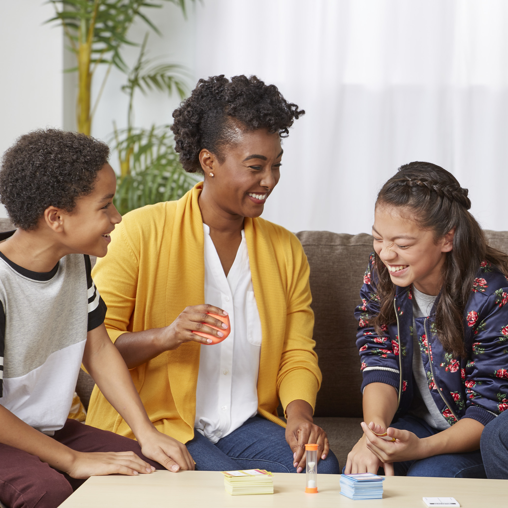 Drie mensen spelen een bordspel, lachen. Een vrouw houdt een oranje voorwerp, timer en kaarten op tafel vast.