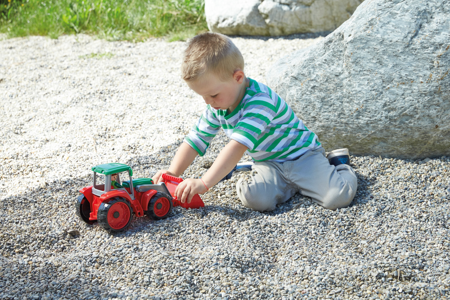 Un niño pequeño juega con un tractor de juguete rojo y verde sobre una superficie de guijarros, cerca de una gran roca y hierba.
