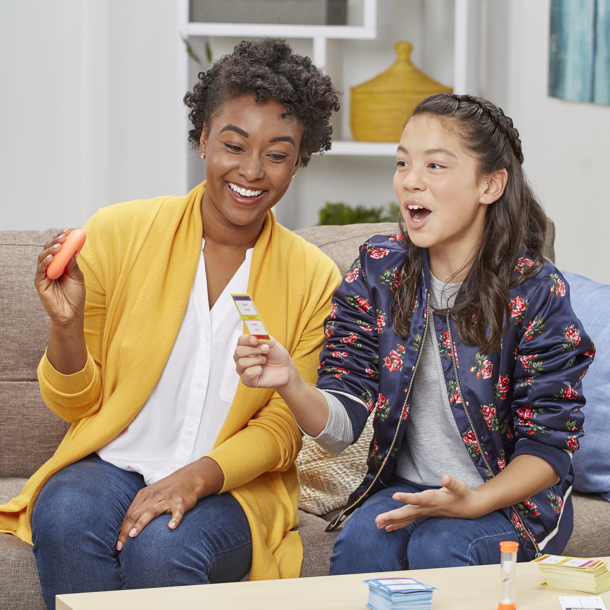 Een vrouw en een meisje spelen een spel. De vrouw houdt een oranje voorwerp vast, het meisje kaarten en heeft haar mond open.