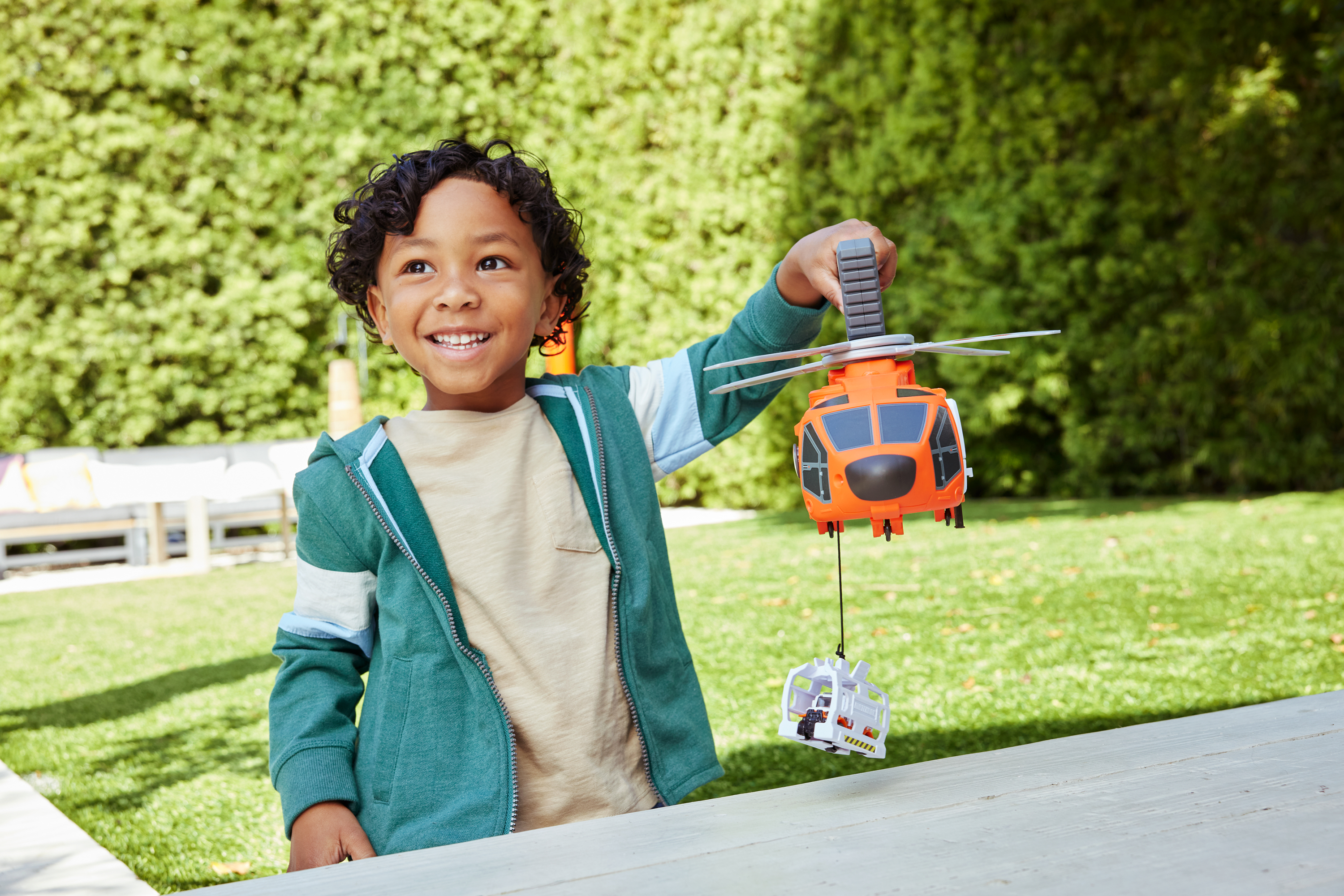 Uśmiechnięty chłopiec trzyma pomarańczowy helikopter. Biały stół i zielona trawa są na pierwszym planie.