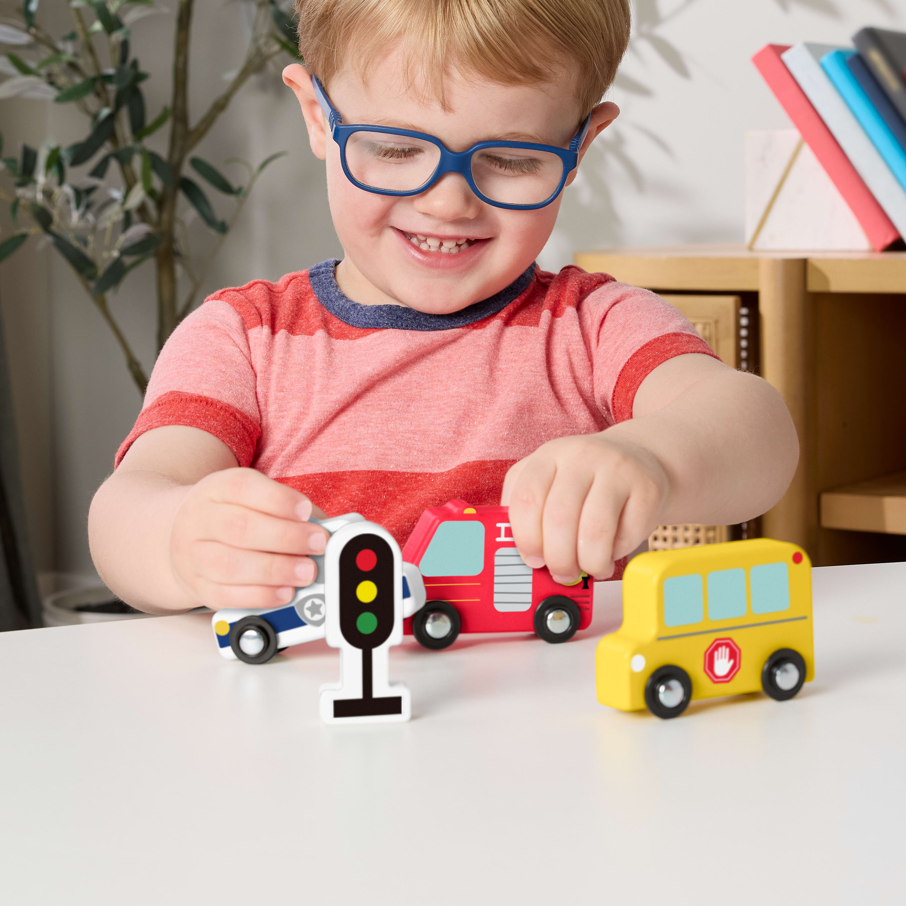 Ein lächelnder Junge mit Brille spielt mit Holzspielzeugautos: Polizeiauto, Feuerwehrauto, Schulbus und Ampel.