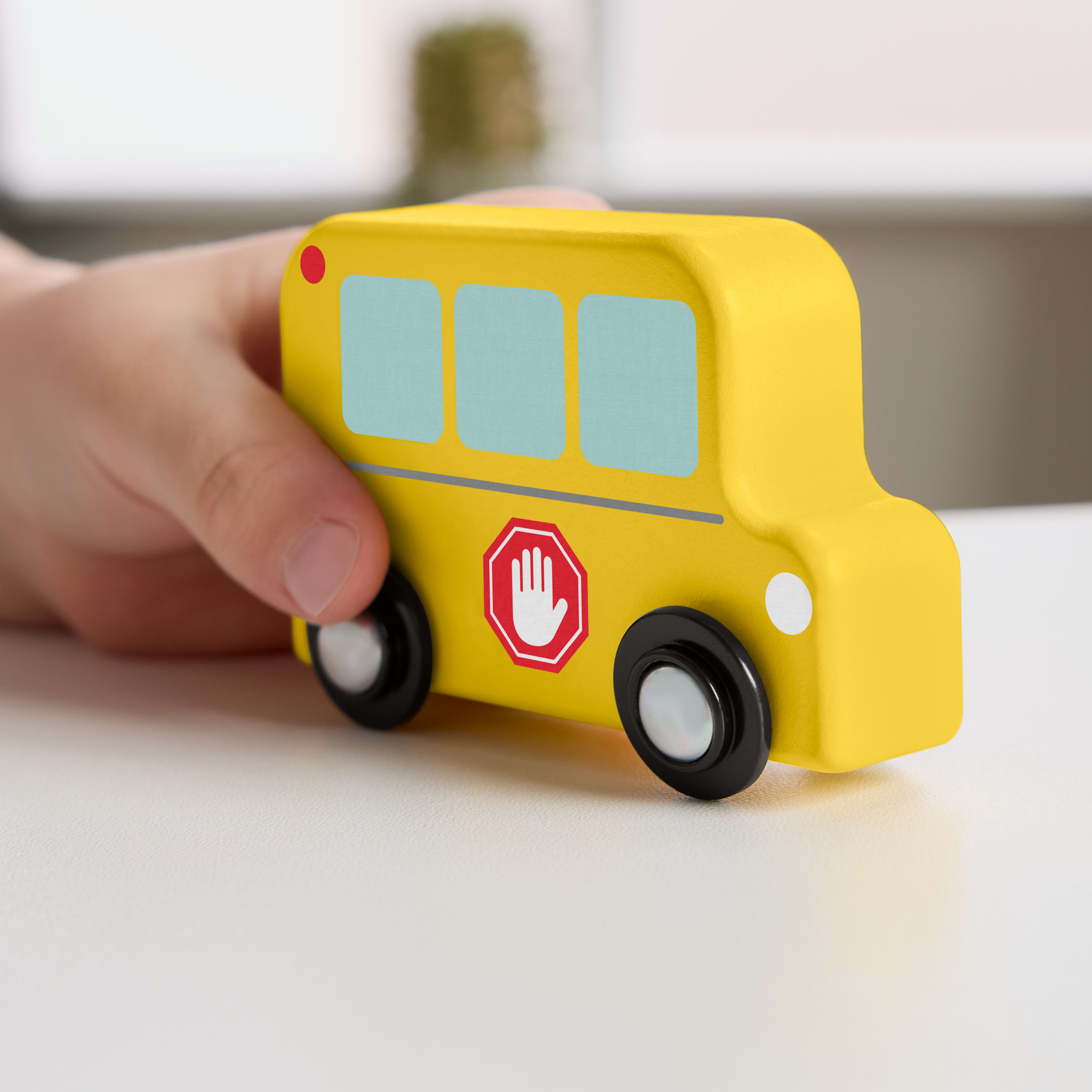 Ein gelber Spielzeug-Schulbus mit schwarzen Rädern, gehalten von einer Hand. Auf der Seite ist ein Stoppschild zu sehen.