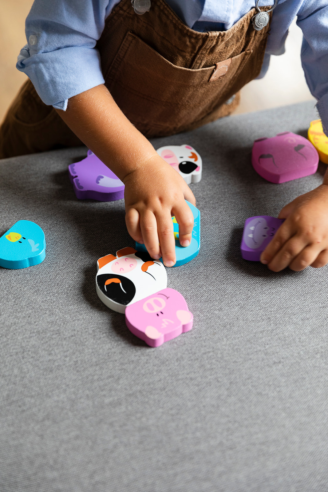 Ręce dziecka bawiące się puzzlami w kształcie zwierząt na szarej powierzchni. Widoczne brązowe ogrodniczki i niebieska koszula.