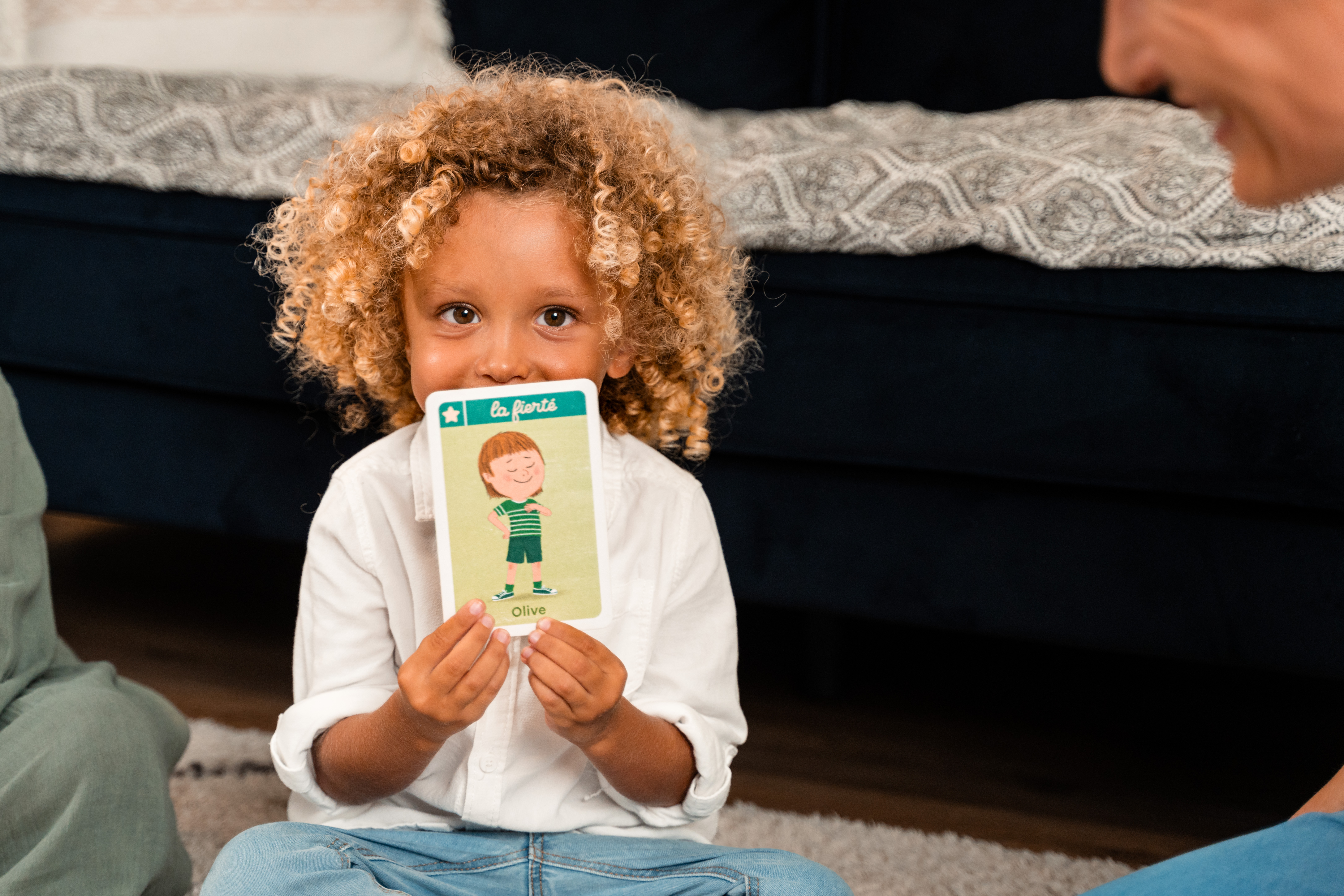 Un bambino con i capelli ricci tiene in mano una carta con scritto 'la fierte'. Il bambino sorride e indossa una camicia bianca.