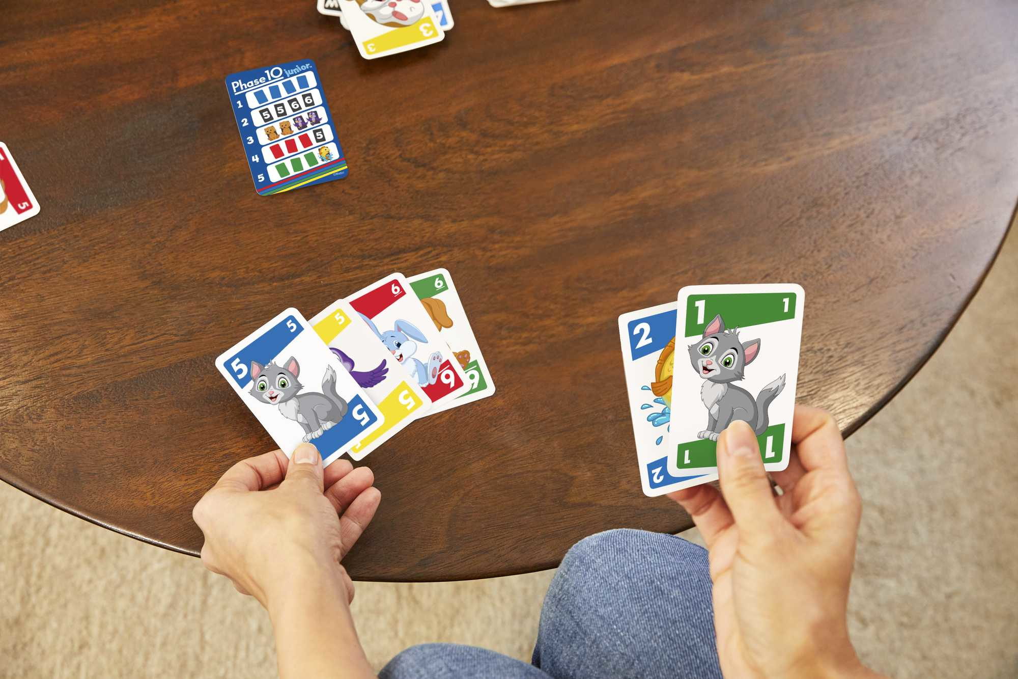 Una persona tiene in mano carte con illustrazioni di gatti. Altre carte e un gioco di carte Phase 10 sono su un tavolo di legno.