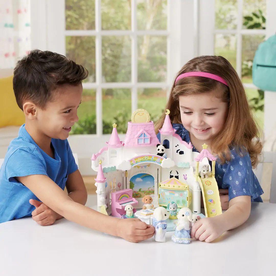 Dos niños juegan con un castillo de juguete. El castillo es blanco y rosa con figuras de juguete. Hay una ventana en el fondo.