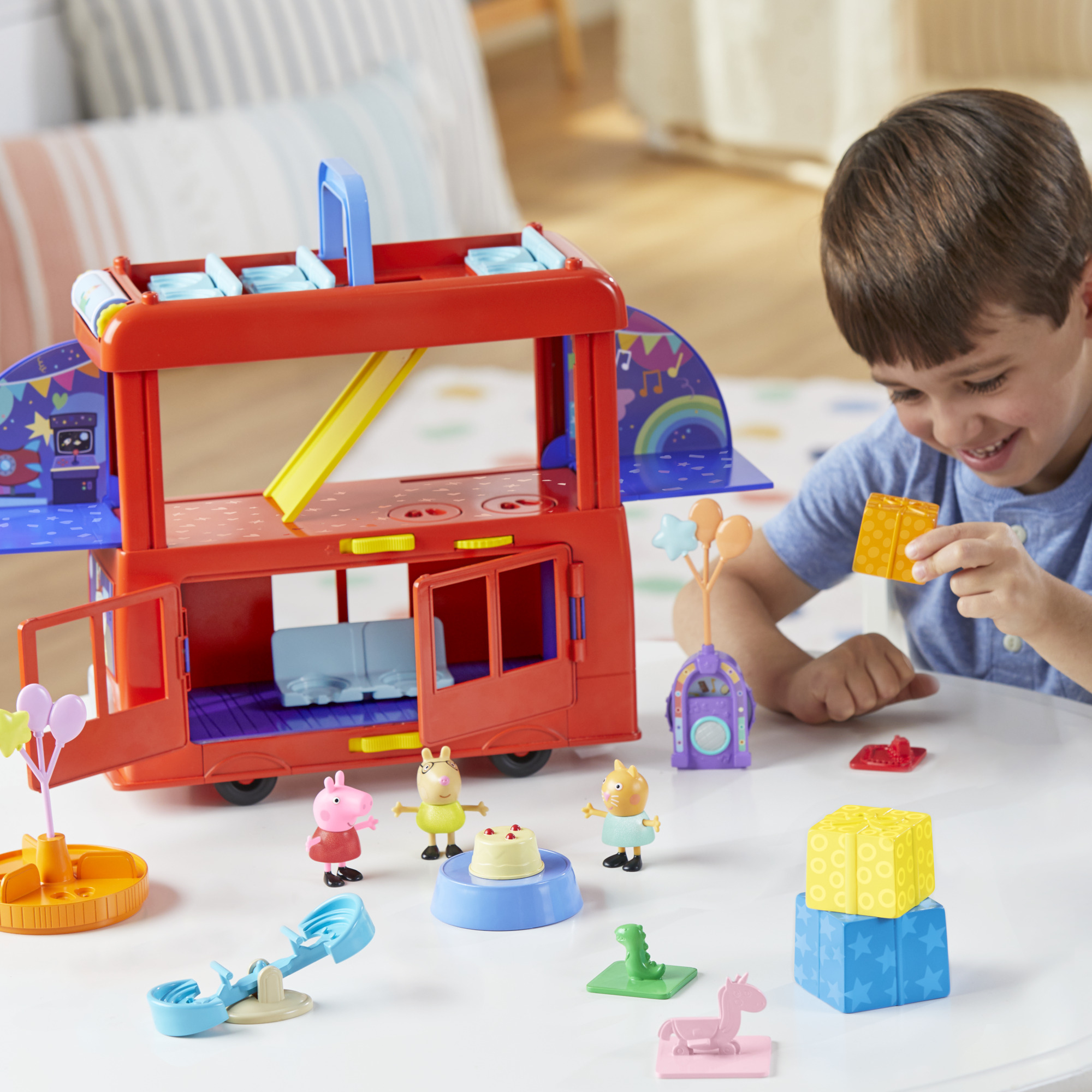 Un niño sonríe, sosteniendo un bloque amarillo. Los juguetes incluyen un autobús rojo, figuritas, pastel y bloques sobre una mesa blanca.