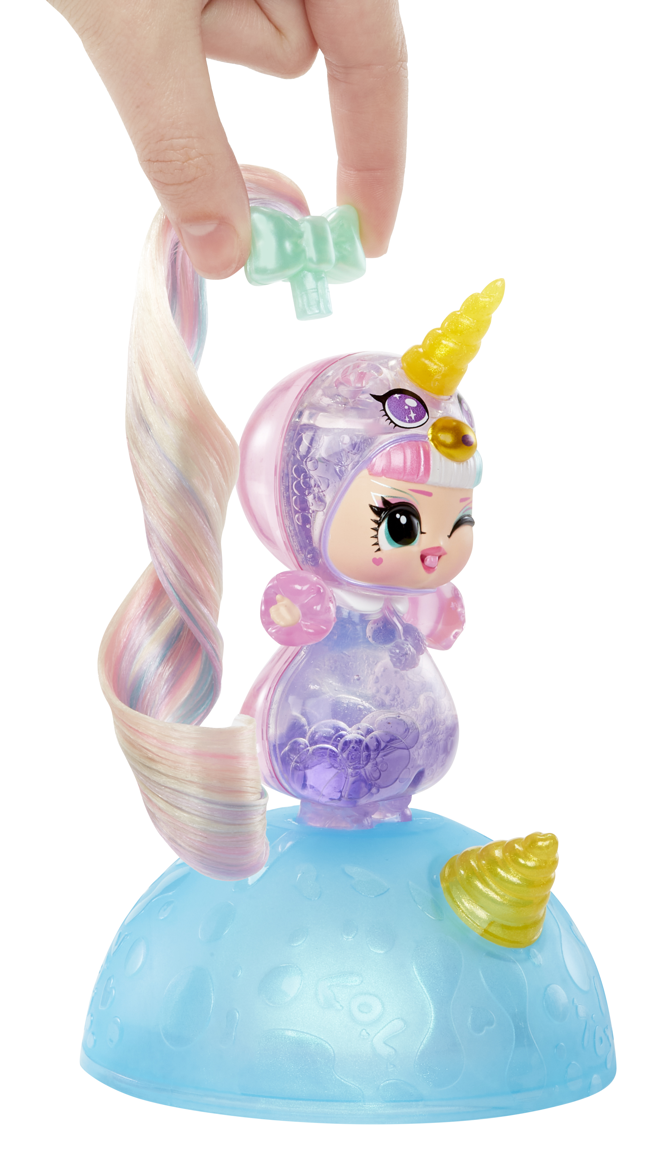 Una mano sostiene una muñeca con un cuerno de unicornio. La muñeca tiene pelo rosa, morado y azul. Está sobre una base azul con un cuerno amarillo.