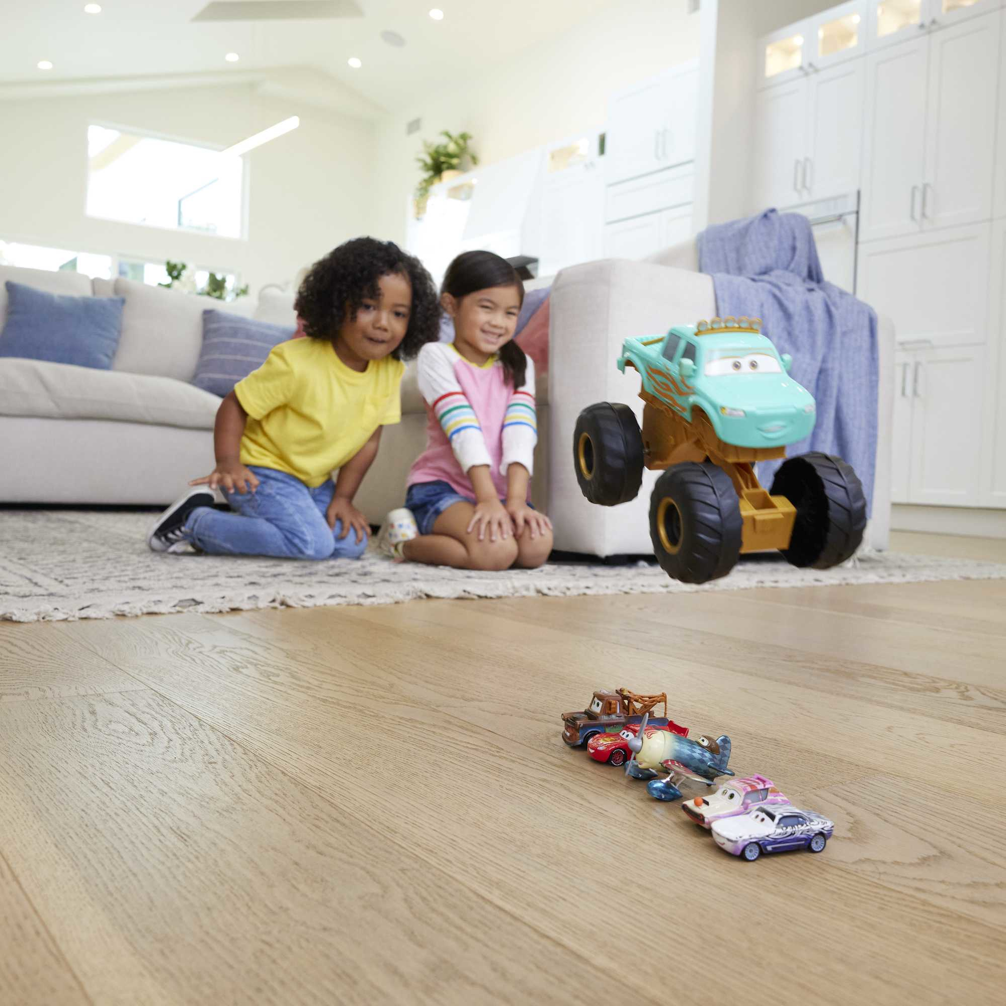 Dwoje dzieci klęczy, patrząc na zabawki samochodowe na podłodze. Duży monster truck unosi się w pobliżu.