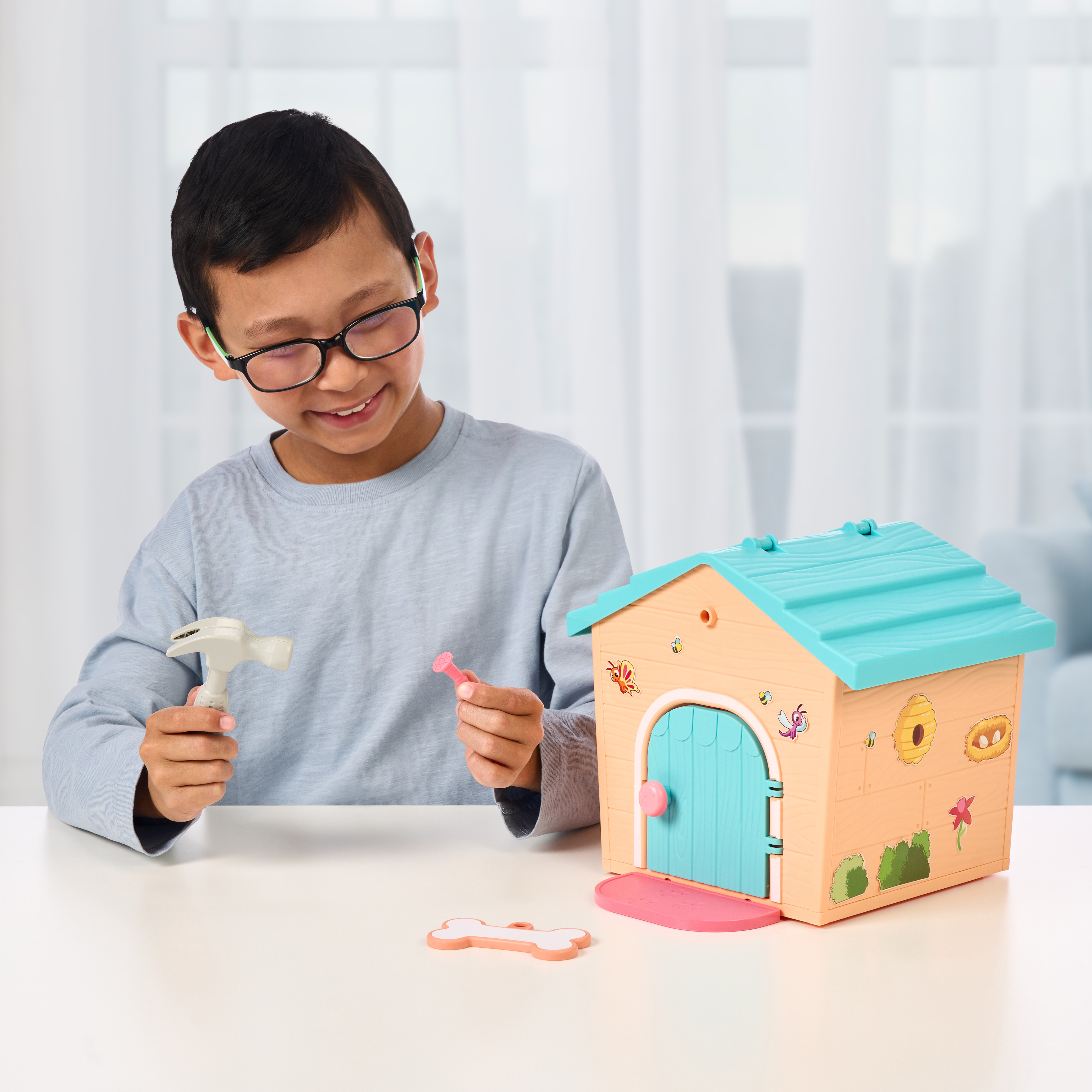 Chłopiec w okularach bawi się zabawnym domkiem dla psa. Trzyma młotek i gwóźdź, uśmiechając się. Dom jest na białym stole.