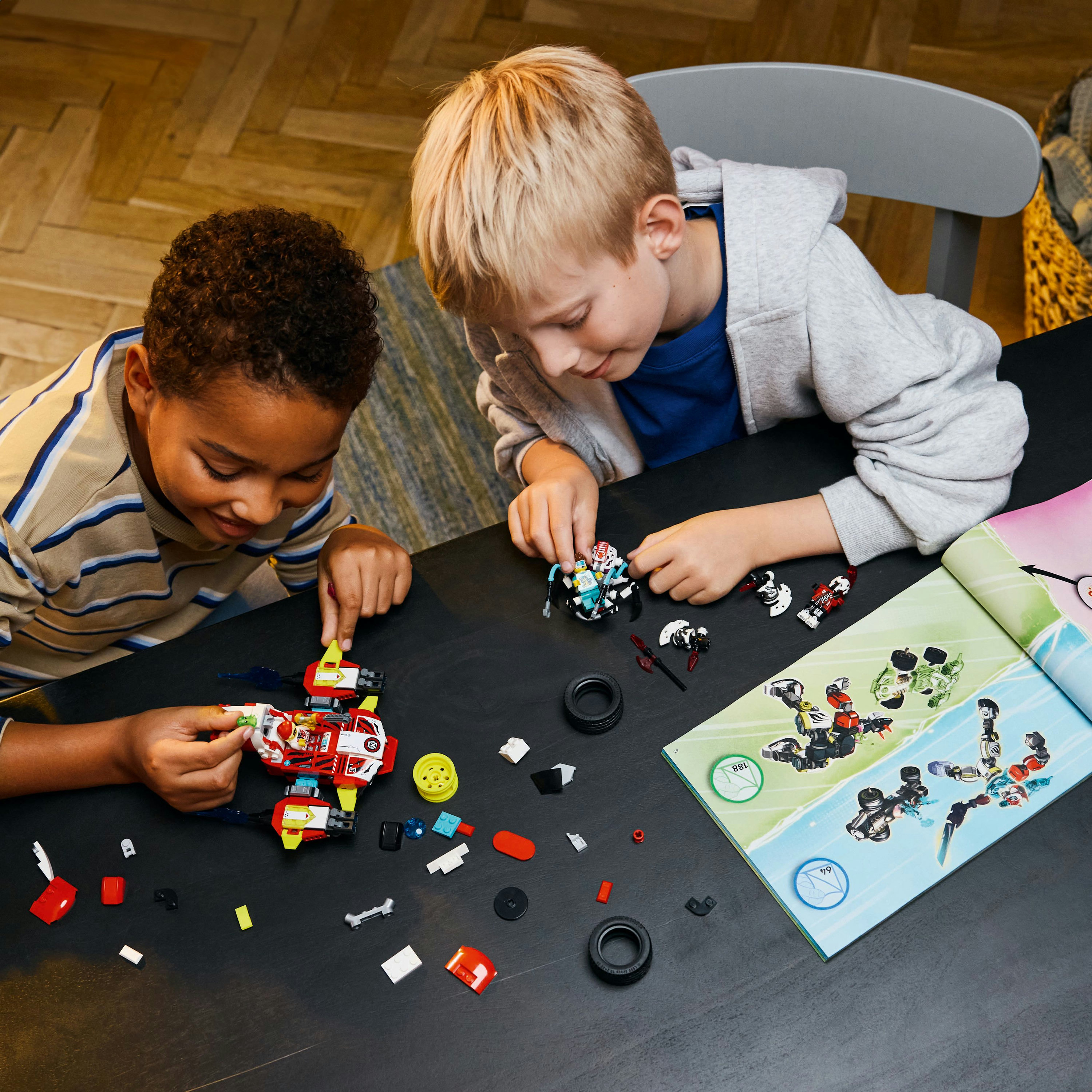 Dwóch chłopców buduje zestawy Lego na czarnym stole. Części są rozrzucone. Jeden nosi niebieską koszulę, drugi w paski.