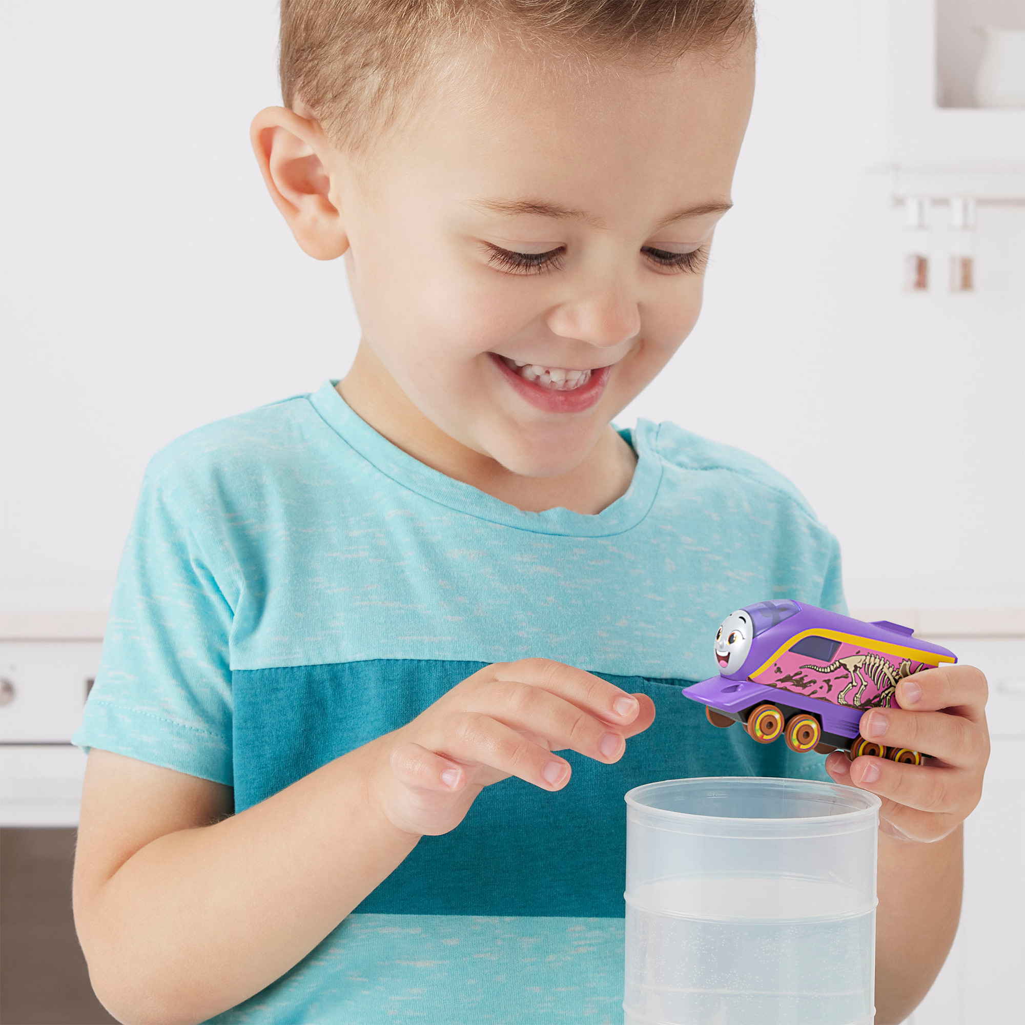 Uśmiechnięty chłopiec trzyma fioletową zabawkę-pociąg. Pociąg ma wzór dinozaura. Z przodu stoi przezroczysty pojemnik.