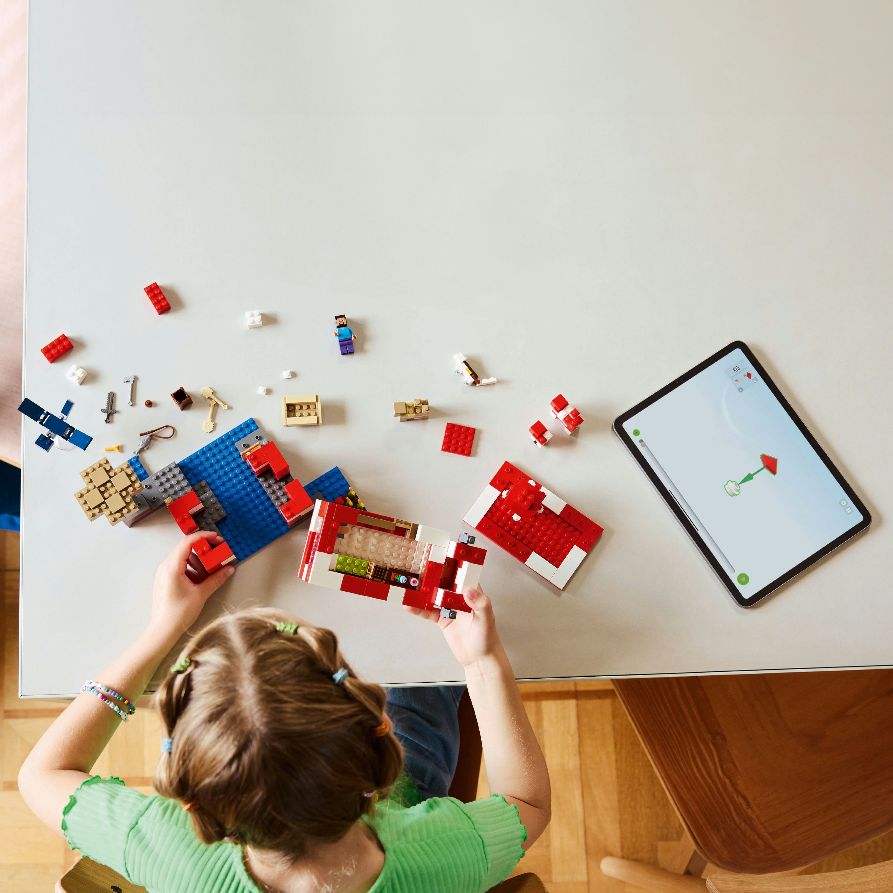 Dziewczynka buduje z klocków Lego na białym stole. iPad wyświetla instrukcje budowy. Różne elementy Lego rozrzucone.