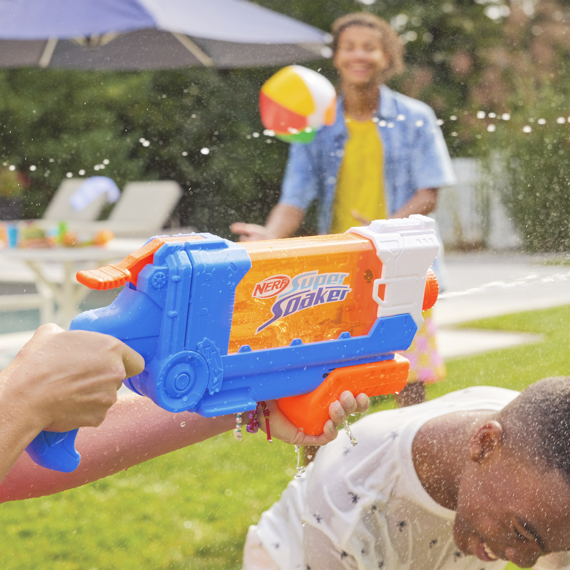 Osoba spryskuje wodą z niebiesko-pomarańczowego Super Soaker inną osobę. Piłka plażowa jest w powietrzu.