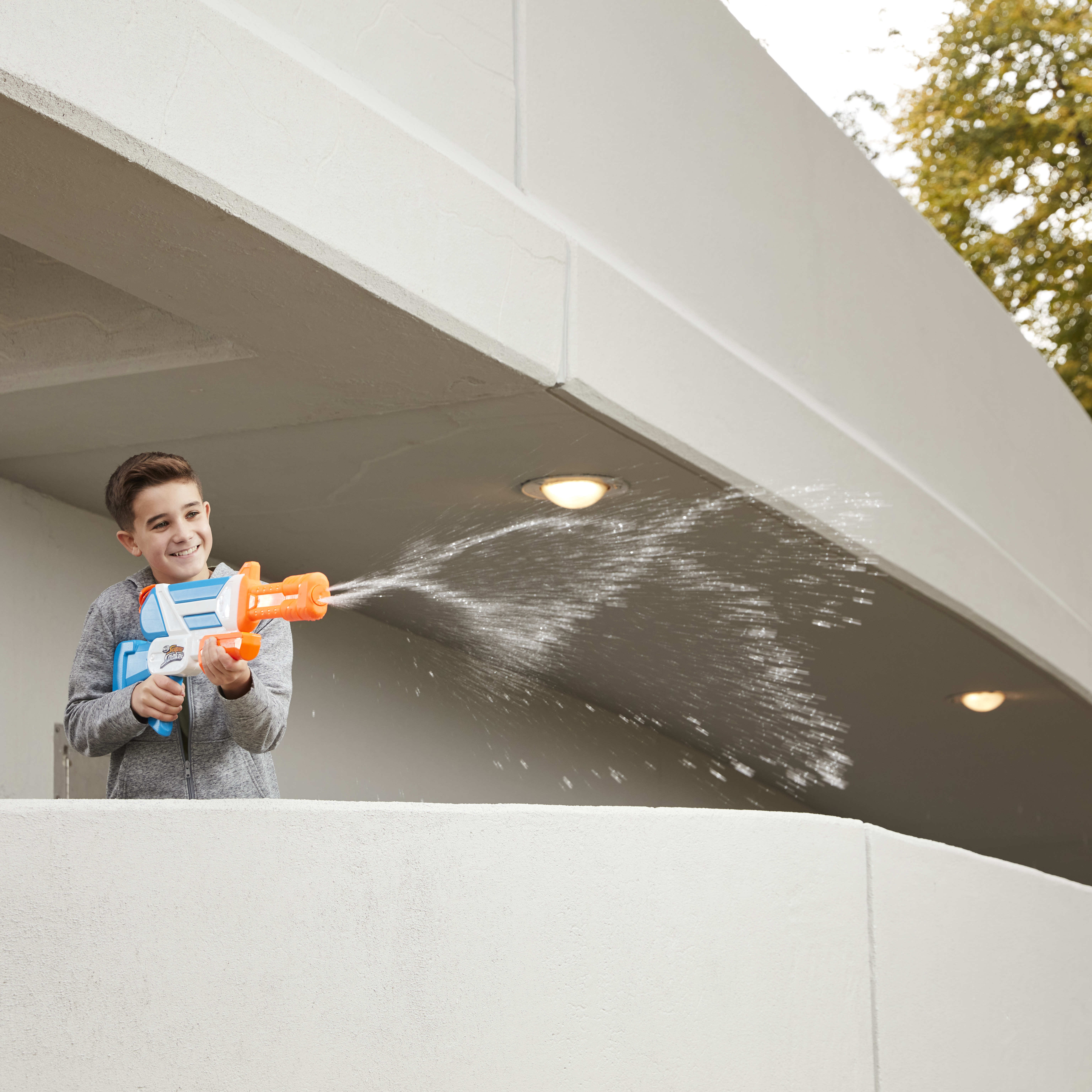 Un niño sonríe, apuntando con una pistola de agua azul y naranja, rociando agua. Una estructura blanca al fondo.