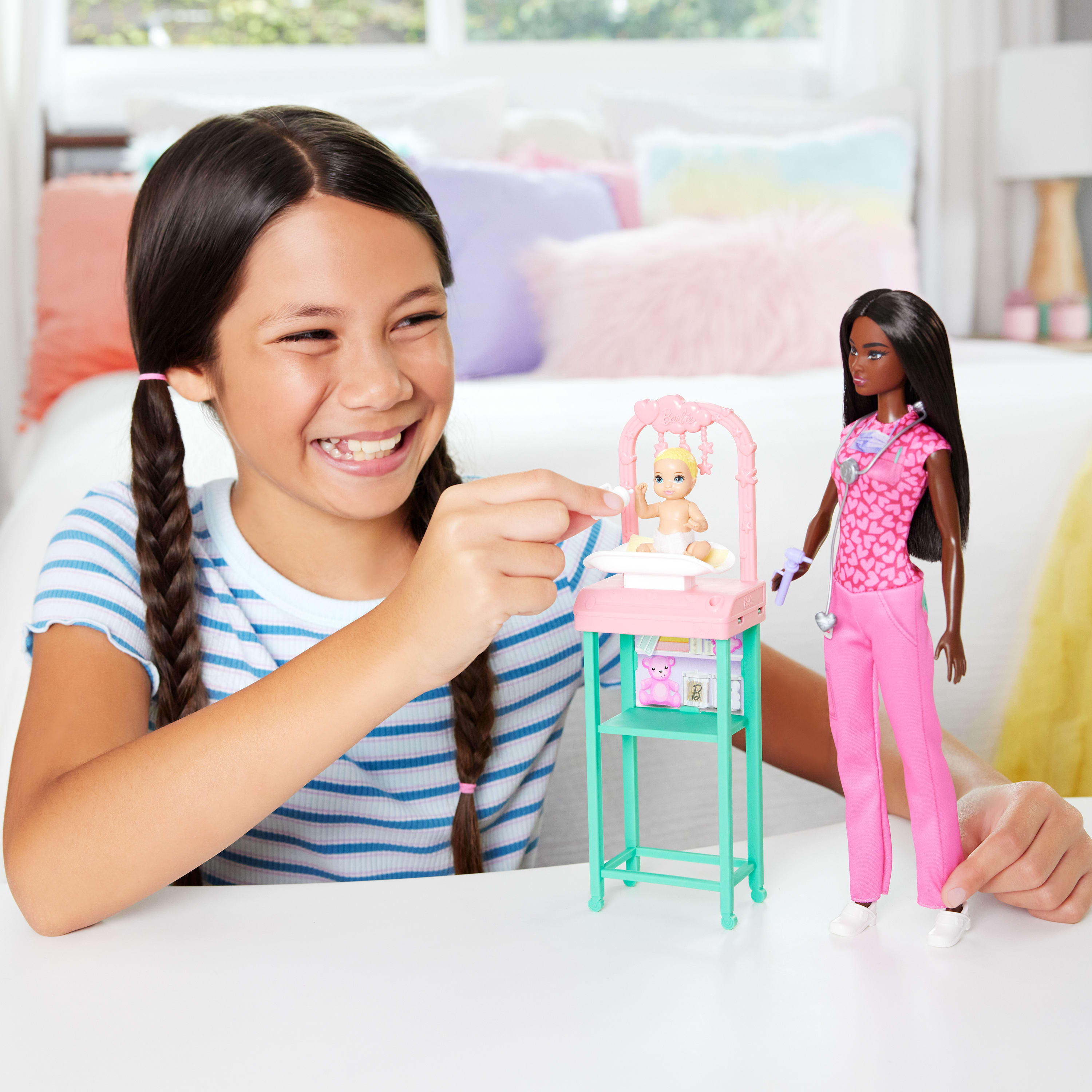 Ein Mädchen lächelt und spielt mit Puppen. Eine Puppe ist Ärztin, die andere ein Baby in einem rosa Spielbereich. Im Hintergrund ist ein Bett.