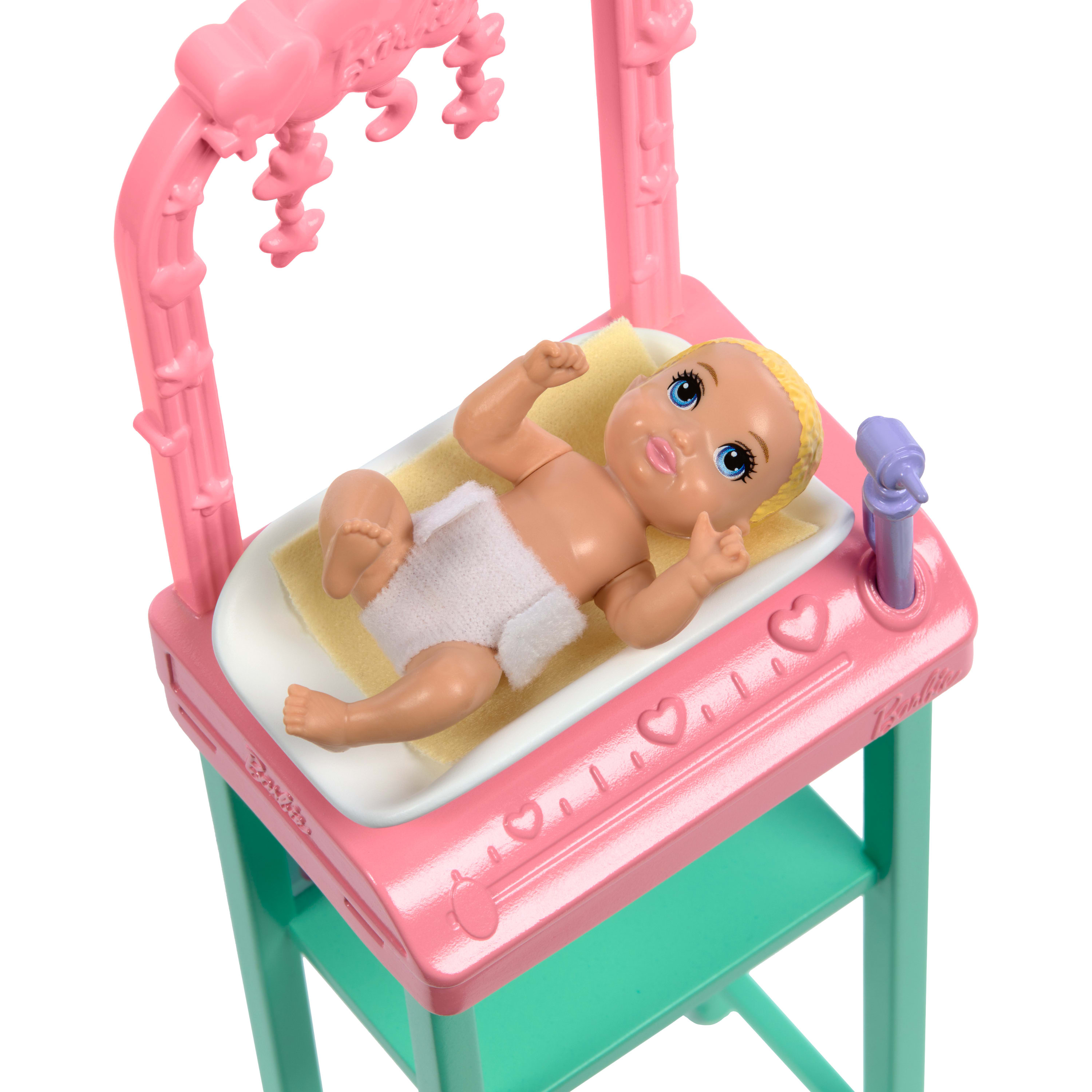 Eine Babypuppe liegt auf einem rosa Wickeltisch. Die Puppe hat blaue Augen und eine Windel. Der Tisch steht auf einem grünen Ständer.