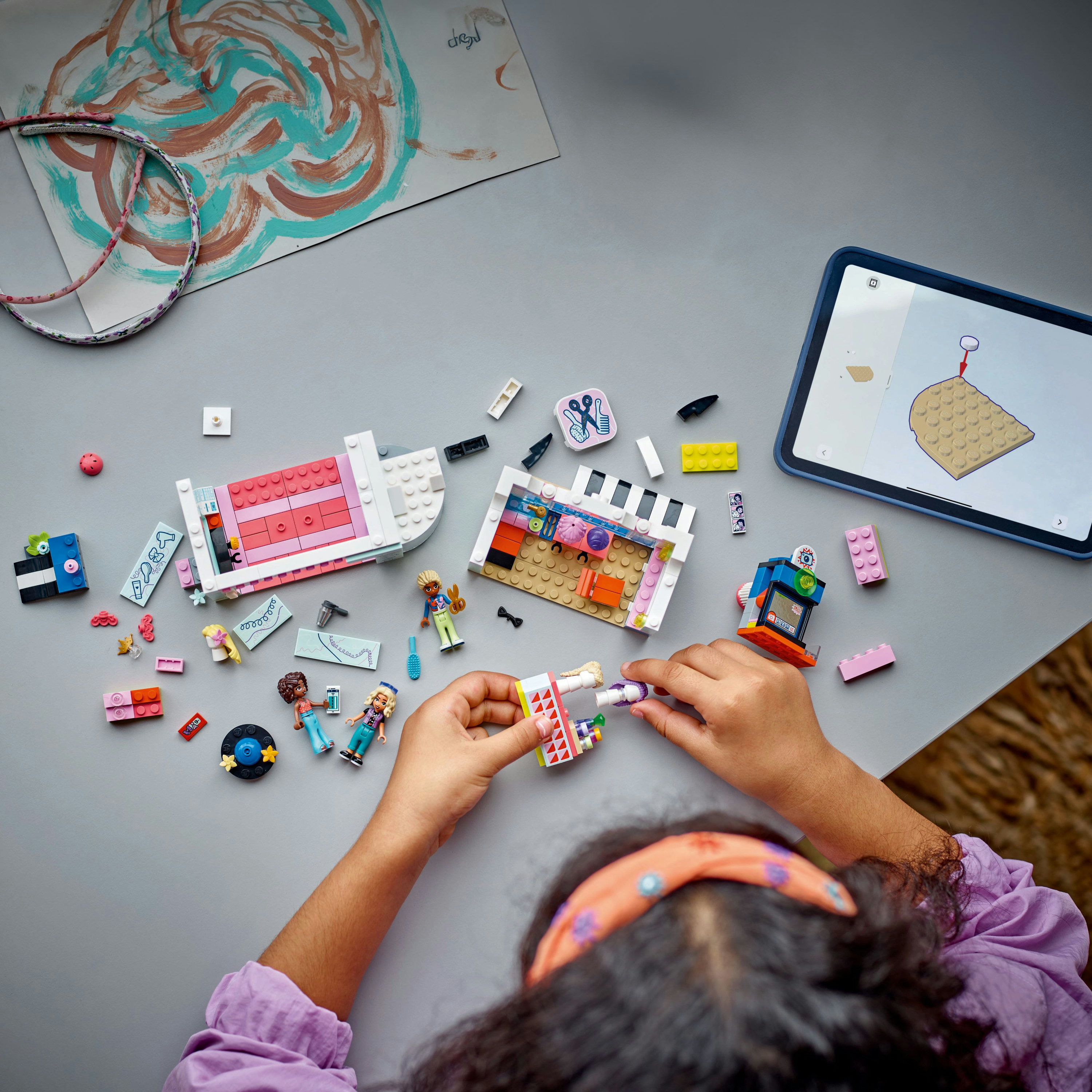 Dziecko buduje zestaw Lego. Scena obejmuje różne elementy Lego, figurki i tablet z instrukcjami.