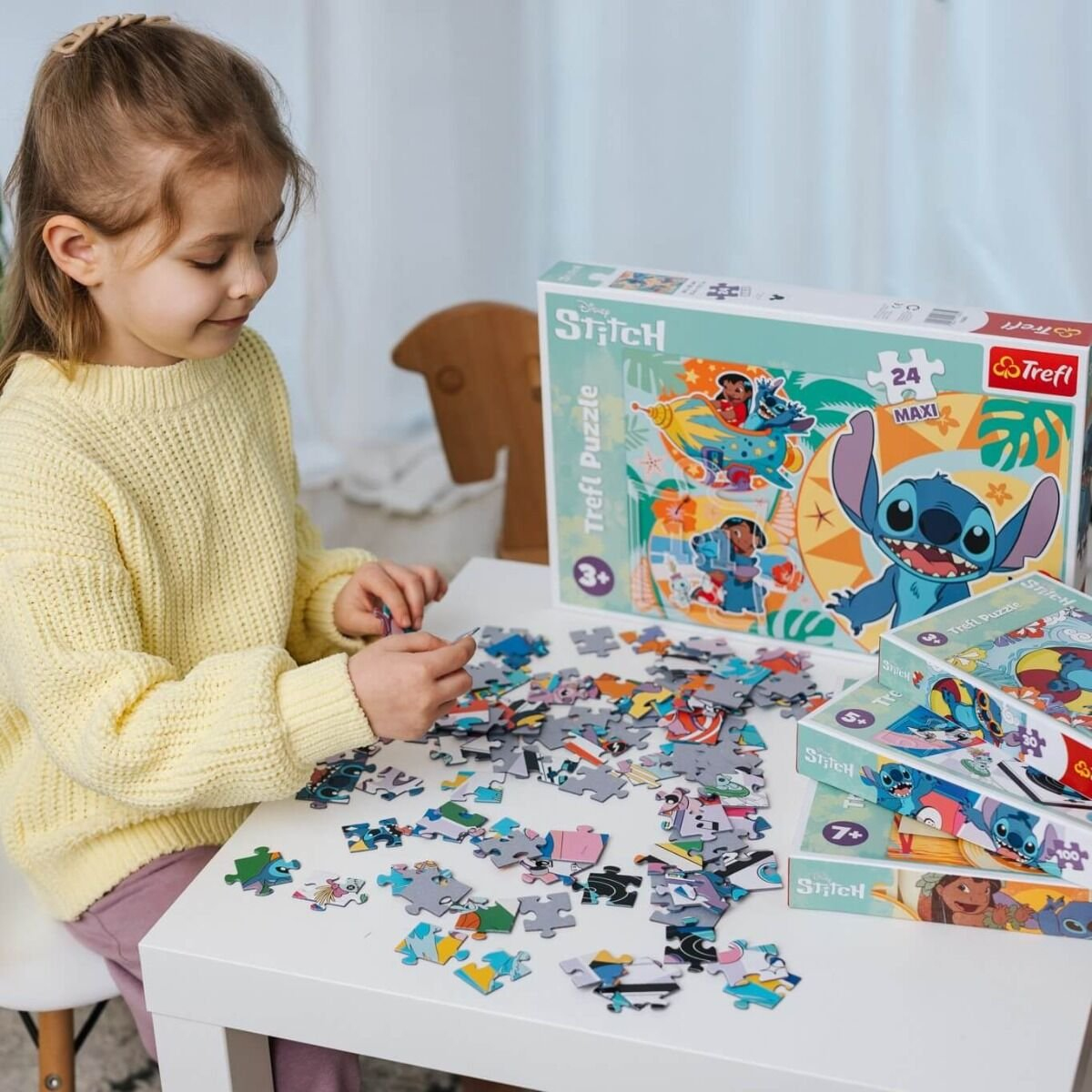 Dziewczyna w żółtym swetrze układa puzzle Stitch na białym stole. Rozrzucone są elementy i pudełka puzzli.