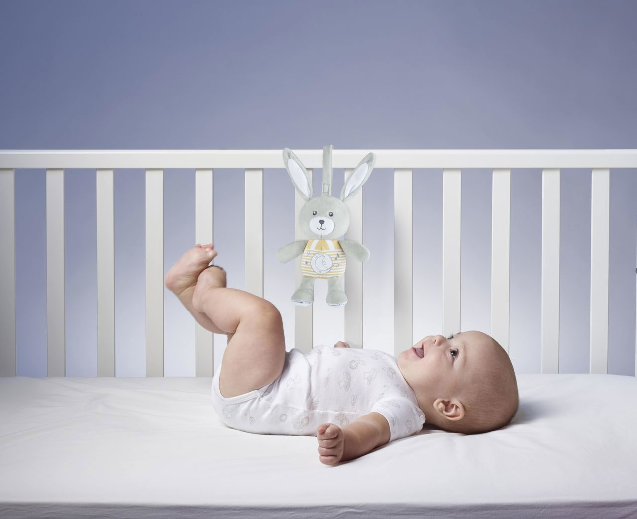 Niemowlę leży w łóżeczku, patrząc na szarą zabawkę królika. Dziecko ma na sobie białe body. Łóżeczko ma białe szczebelki.