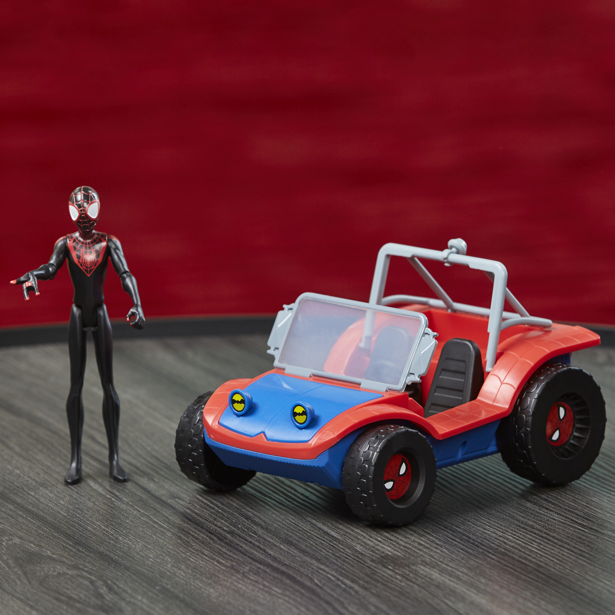 La figura de Spider-Man está junto a un coche de juguete rojo, azul y negro sobre un fondo rojo, en una superficie similar a la madera.