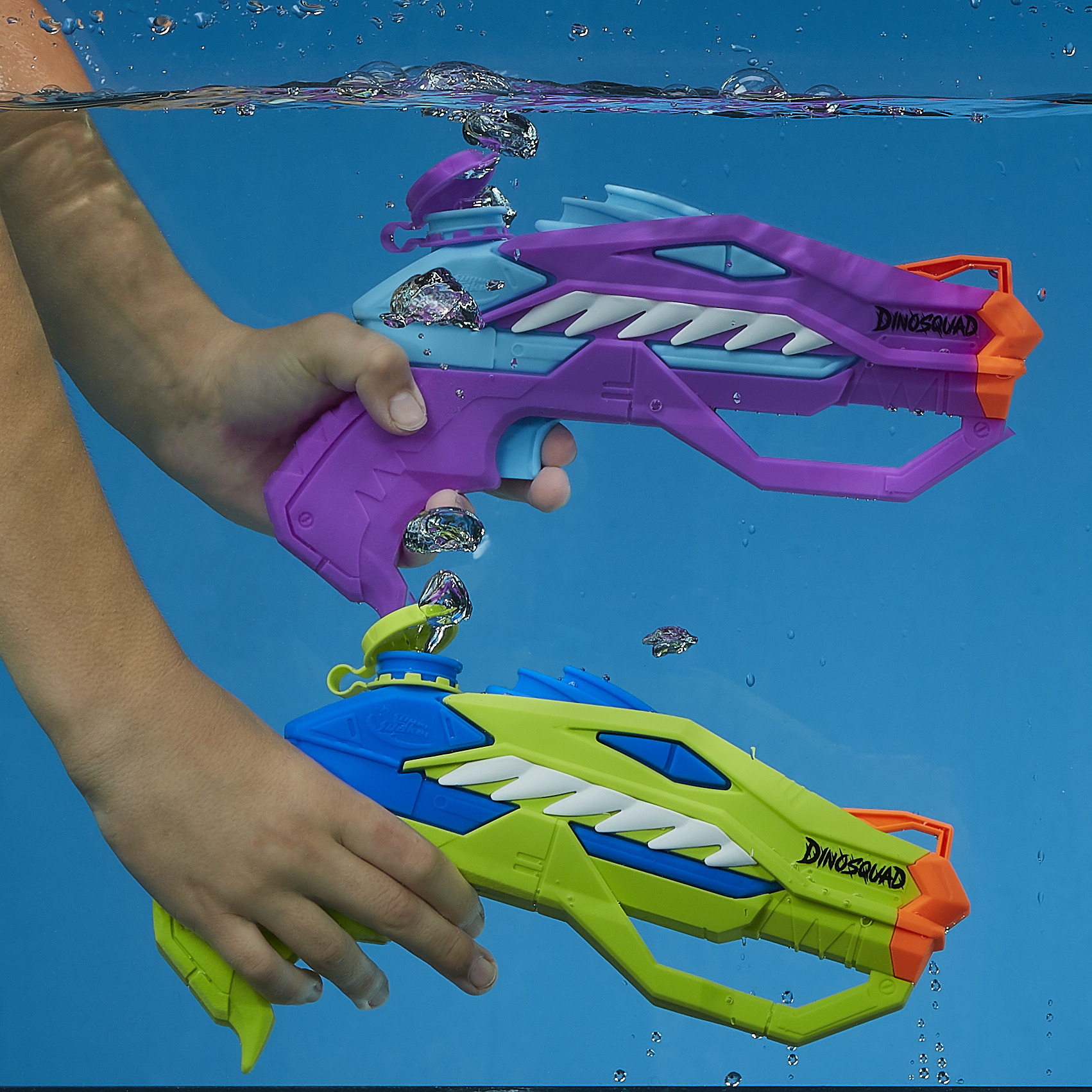 Dwa pistolety na wodę z motywem dinozaurów są trzymane pod wodą. Pistolety są fioletowe i zielone, z białymi zębami. Widoczne są bąbelki.
