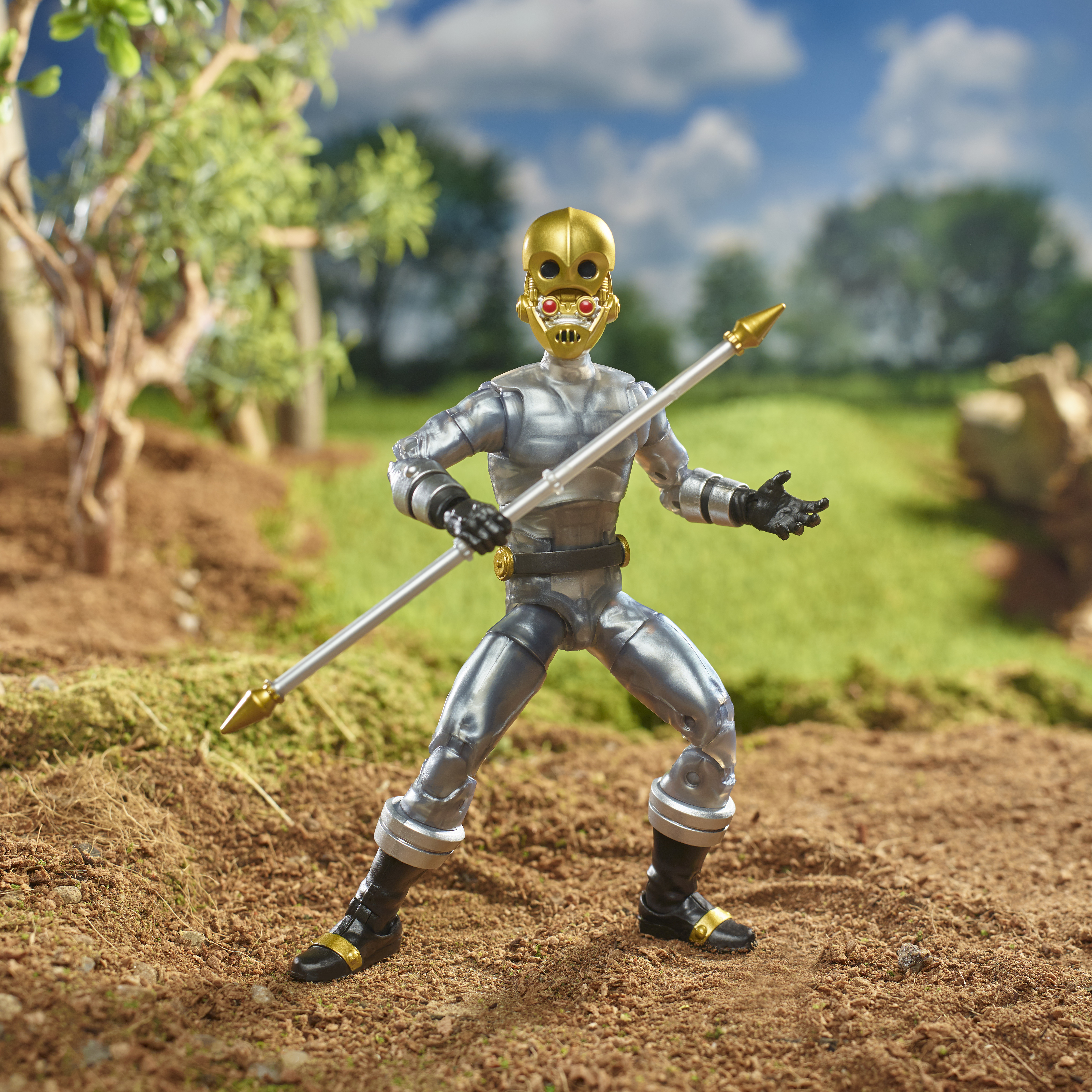 Action figure di un personaggio con maschera d'oro che tiene una lancia, in posa su una superficie marrone con sfondo verde.