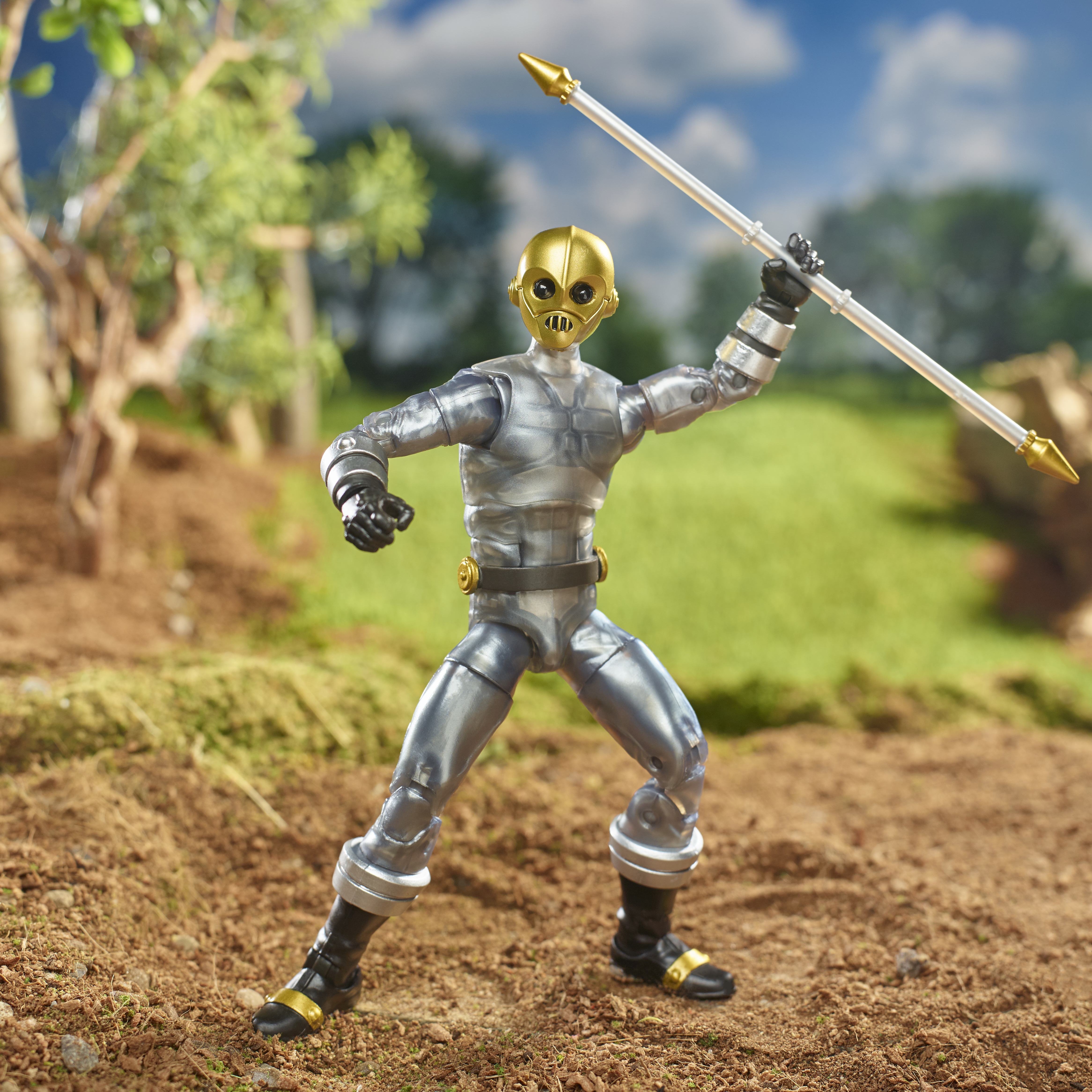 Una figura d'argento con casco e lancia d'oro è in piedi sulla terra. Lo sfondo ha erba, alberi e cielo blu.