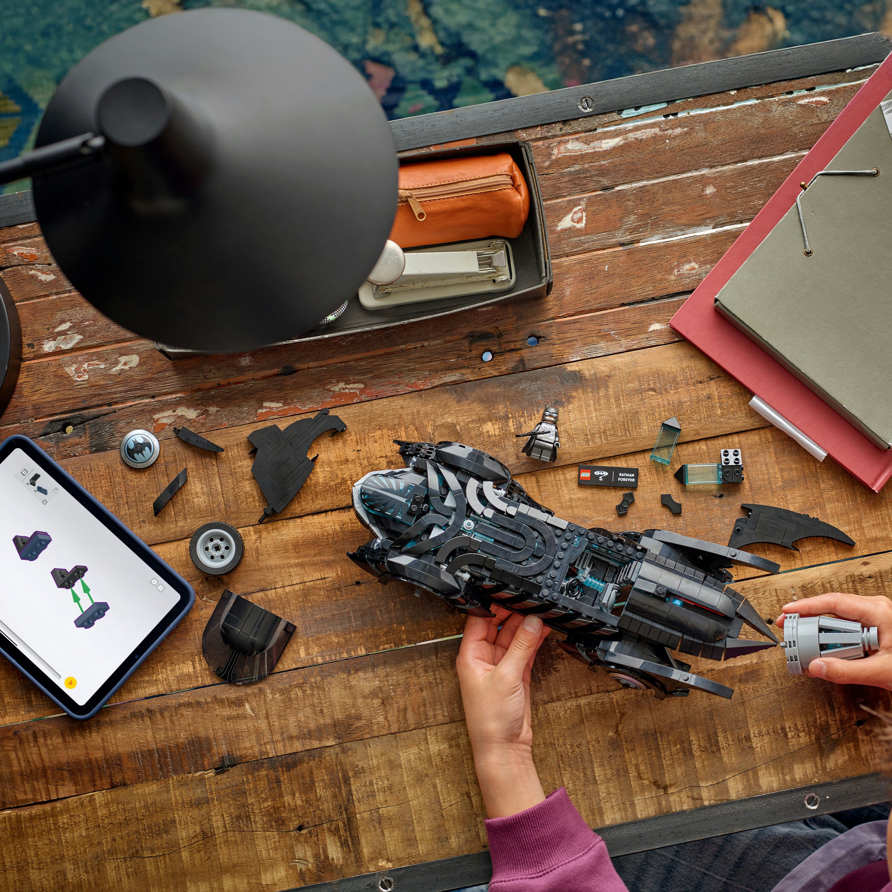 Osoba składająca czarny Batmobil Lego na drewnianym stole. Widoczne części i instrukcje. Lampa u góry.