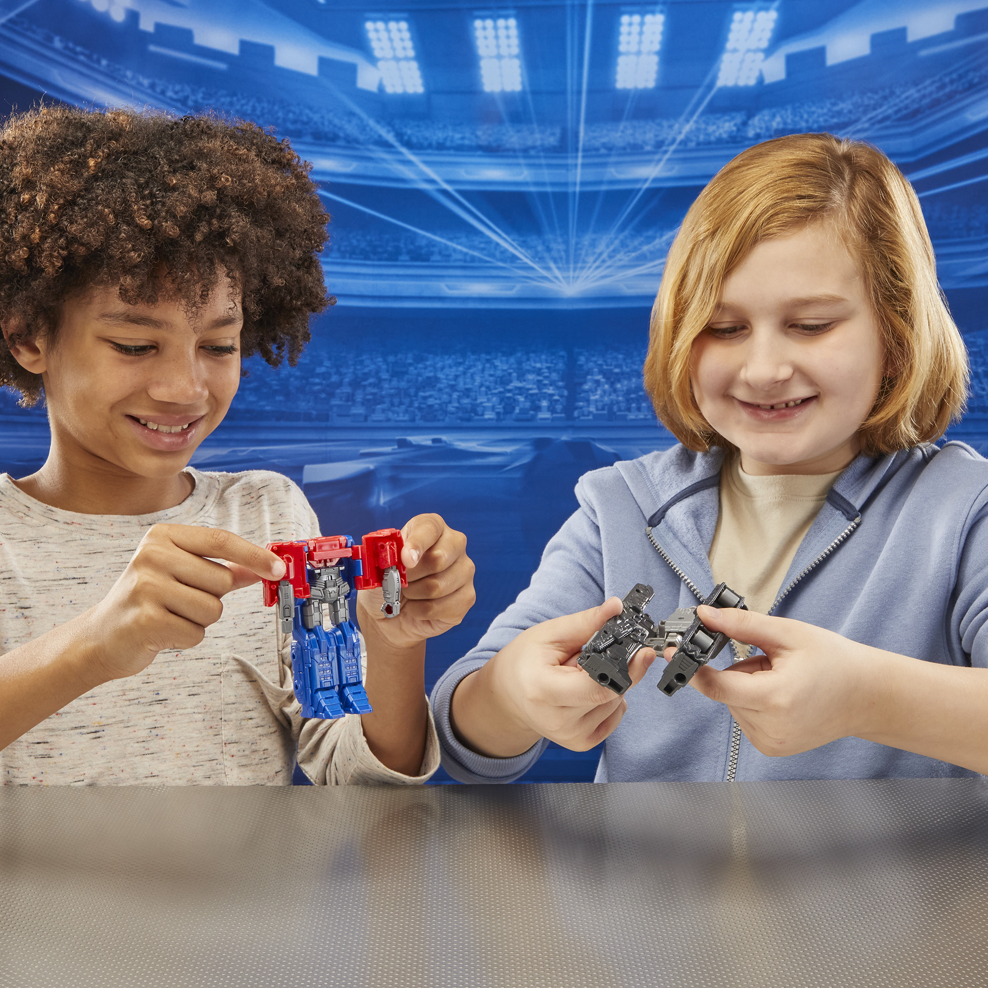 Dos niños sonríen mientras sostienen juguetes Transformers. Uno es rojo y azul, el otro gris, contra un fondo de estadio.