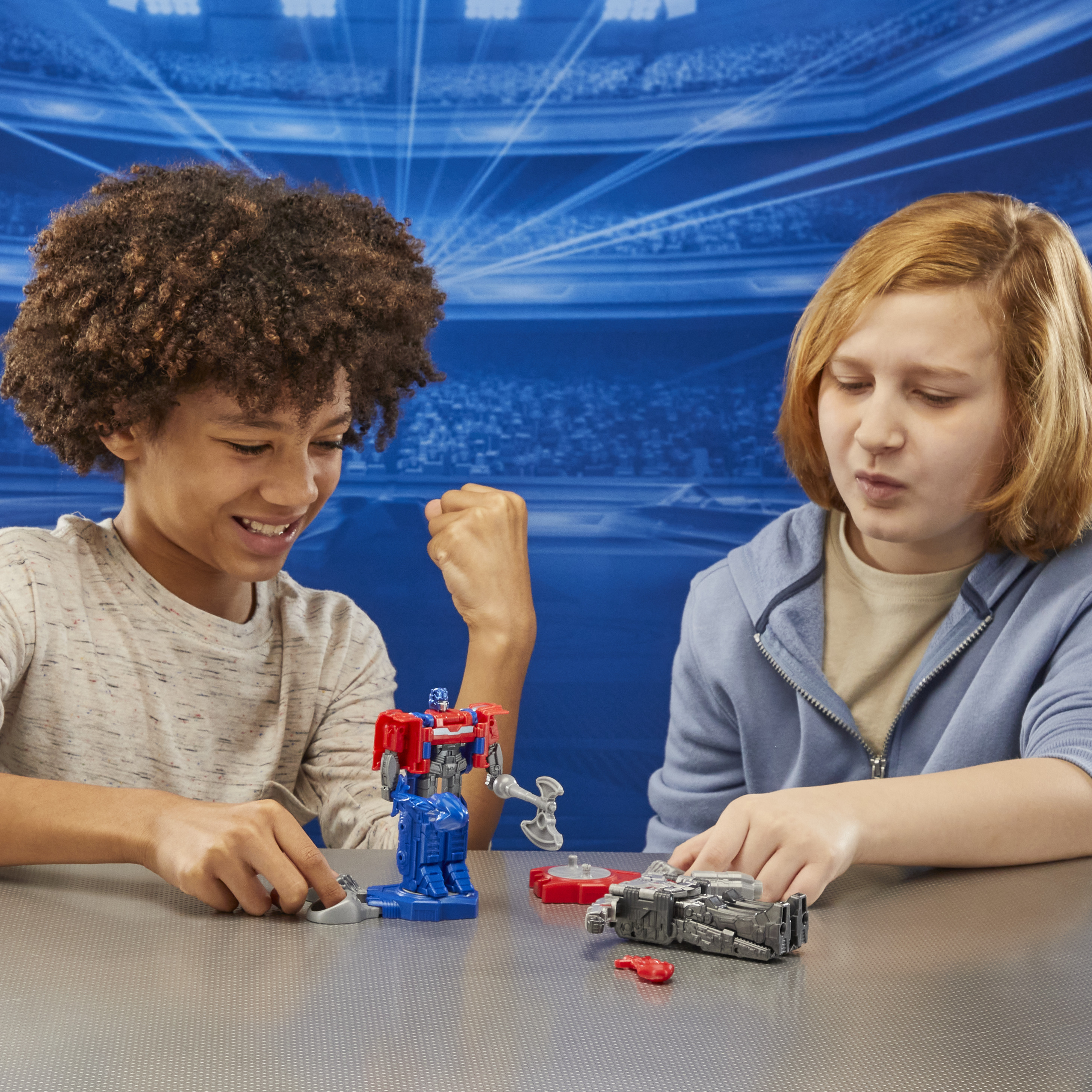 Dos niños juegan con juguetes Transformers. Uno sonríe, el otro parece concentrado. Fondo azul.