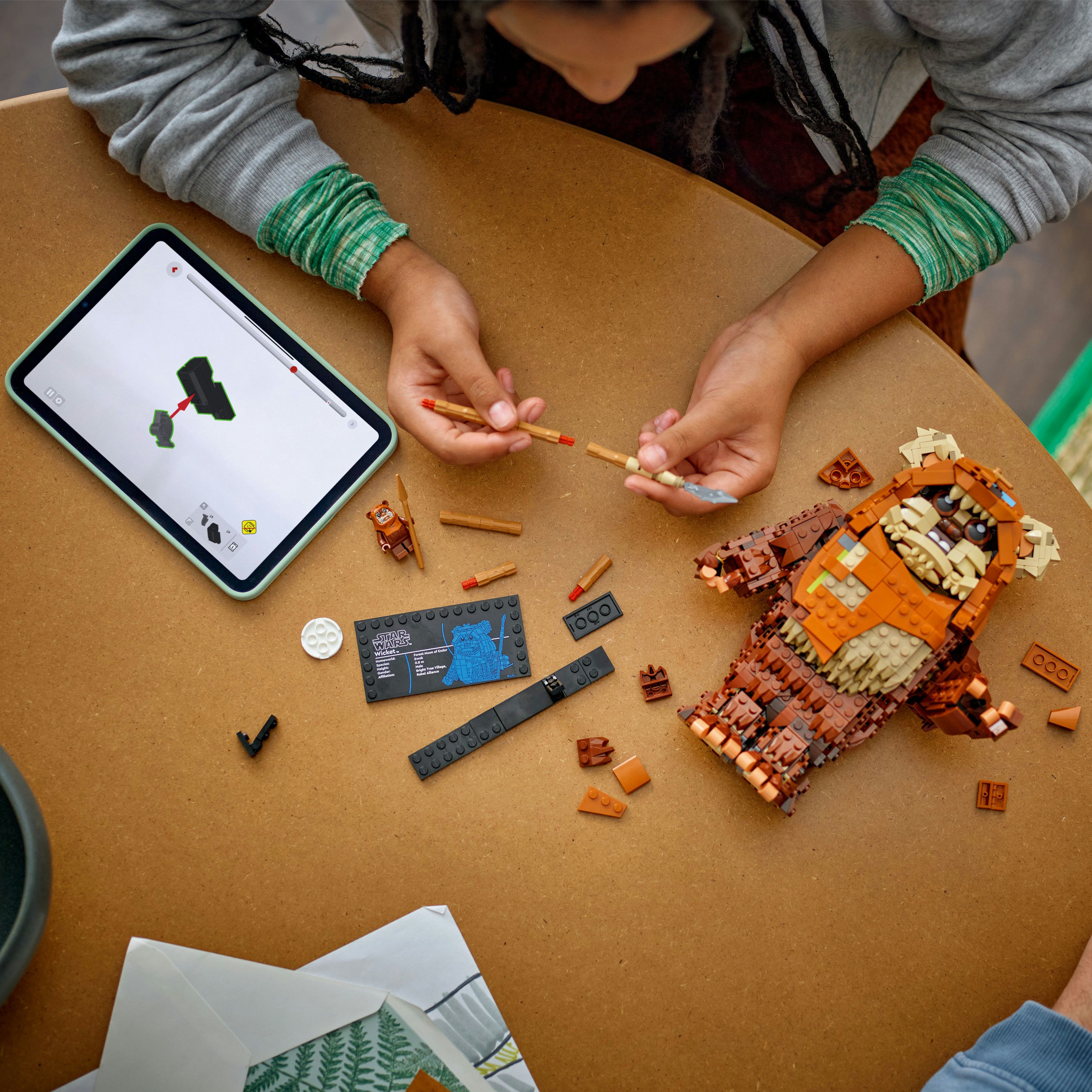 Osoba montuje figurkę Lego Ewok z Gwiezdnych Wojen na brązowym stole. Instrukcje znajdują się na tablecie.