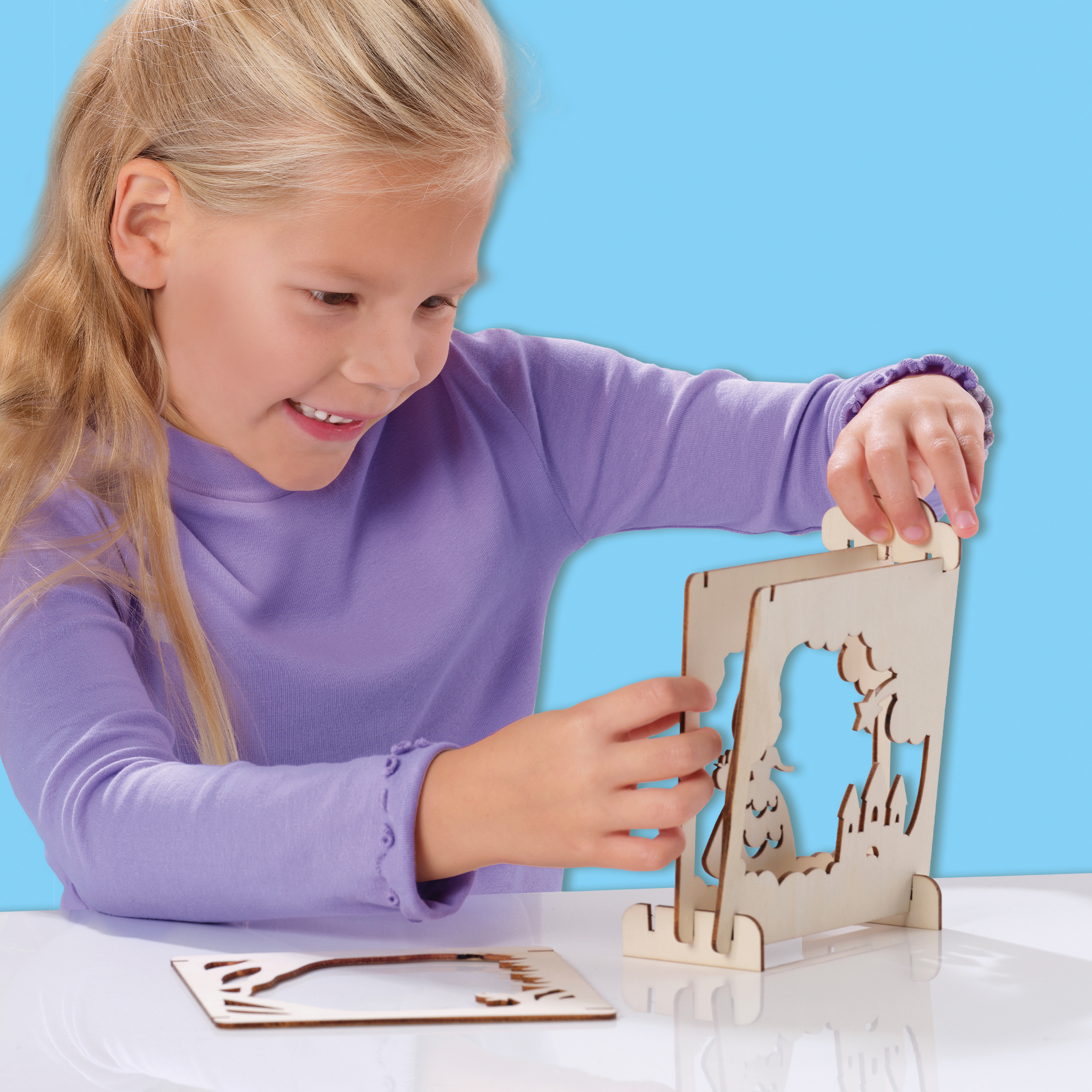 Dziewczyna z blond włosami składa drewnianą zabawkę. Zabawka przedstawia scenę z drzewami i zamkiem.