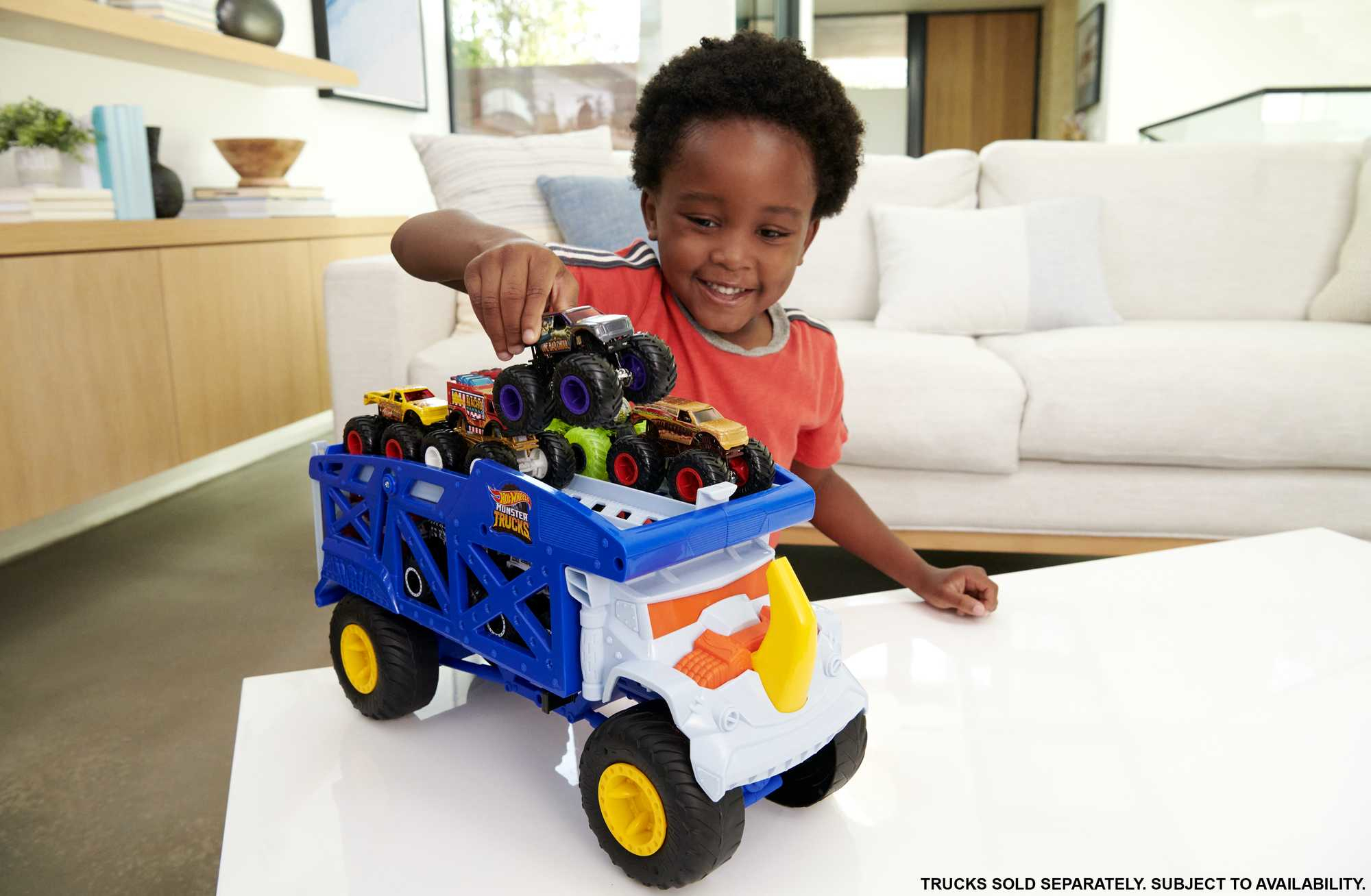 Uśmiechnięty chłopiec umieszcza zabawkę monster truck na niebiesko-białym transporterze. Na transporterze znajdują się inne ciężarówki. Biała kanapa w tle.