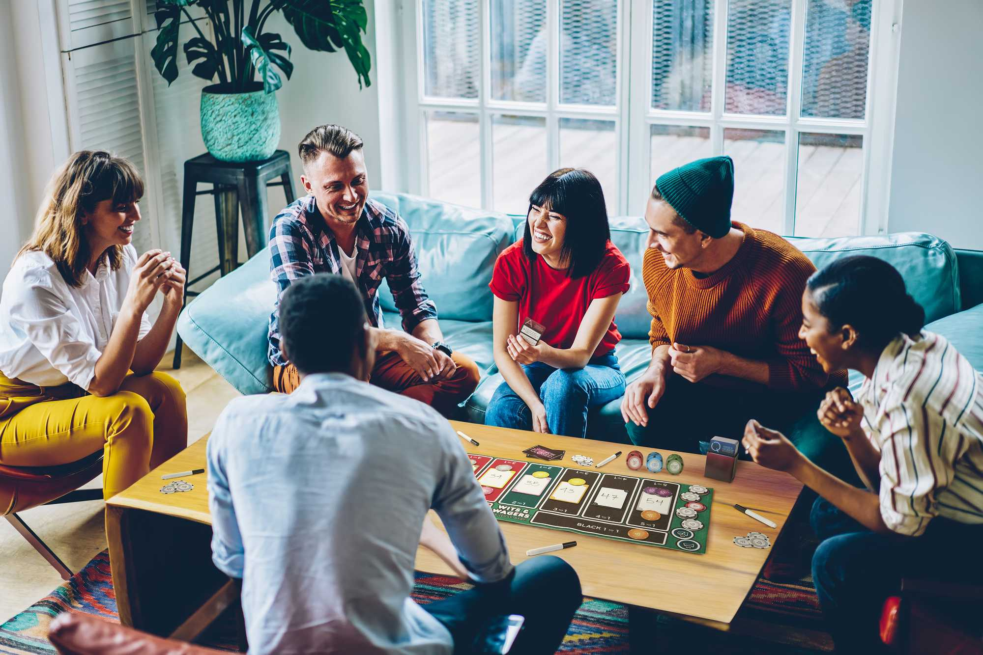 Eine Gruppe von Freunden spielt ein Brettspiel. Sie sitzen um einen Tisch, lächeln und lachen. Der Raum hat natürliches Licht.