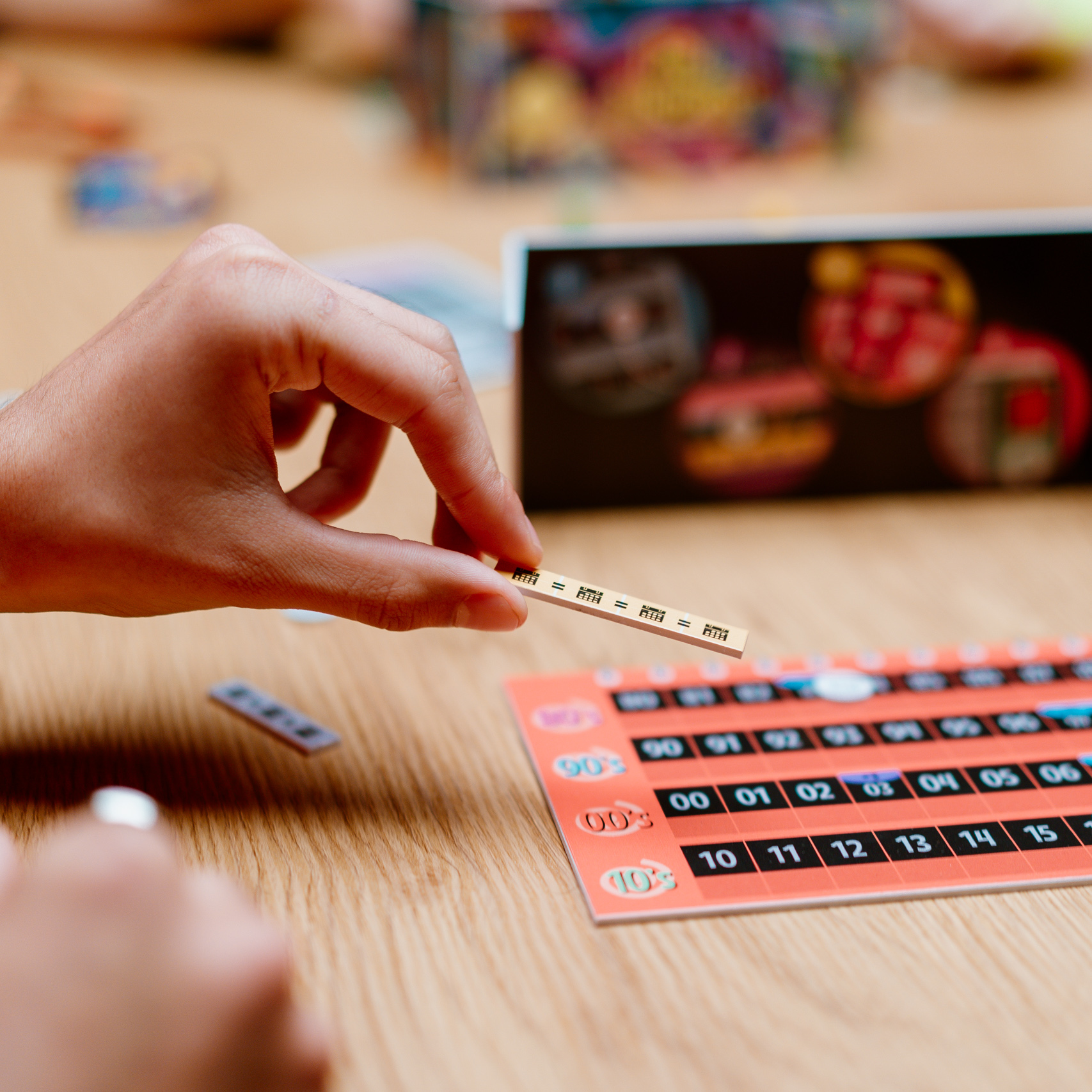 Eine Hand hält ein Holzspielstück über einem Brett. Das Brett ist orange mit Zahlen. Eine andere Hand ist sichtbar.