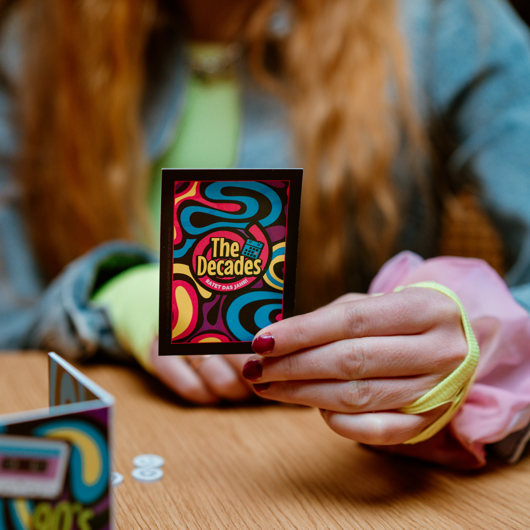 Eine Person hält eine Spielkarte. Die Karte sagt 'The Decades' mit bunten Designs. Im Hintergrund ist eine Spielbox zu sehen.