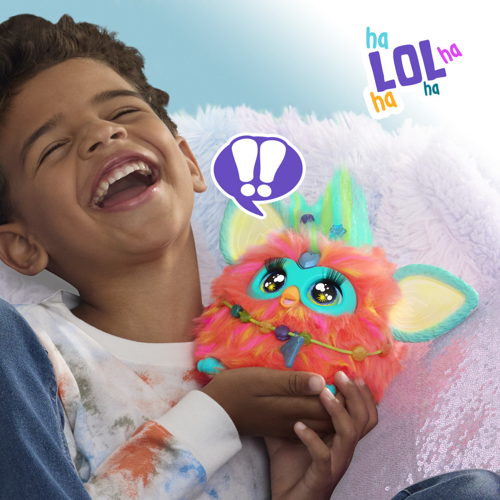 Chłopiec śmieje się, trzymając kolorową zabawkę Furby. Zabawka jest pomarańczowa, turkusowa i żółta. Dymki i 'LOL' w tle.